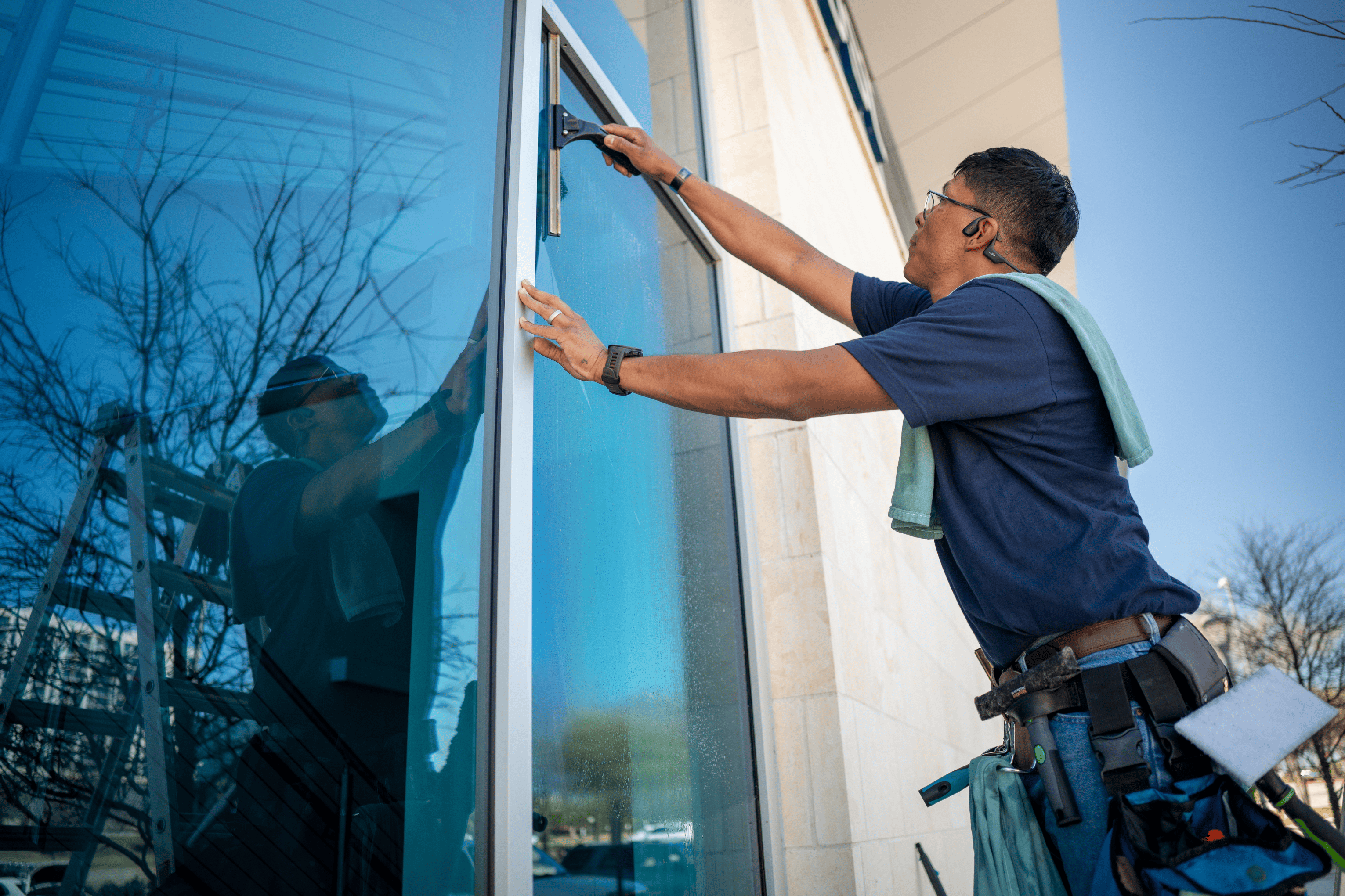 A professional cleaner uses a squeegee to clean a large glass pane on a building exterior under a clear blue sky.