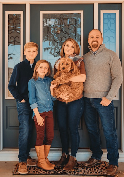 Family of five poses in front of their blue door, including a dog. Smiling faces, casual attire.