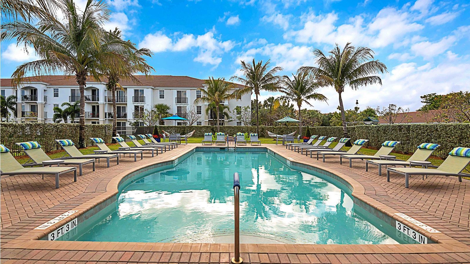 A large swimming pool surrounded by chairs and palm trees in front of a building.
