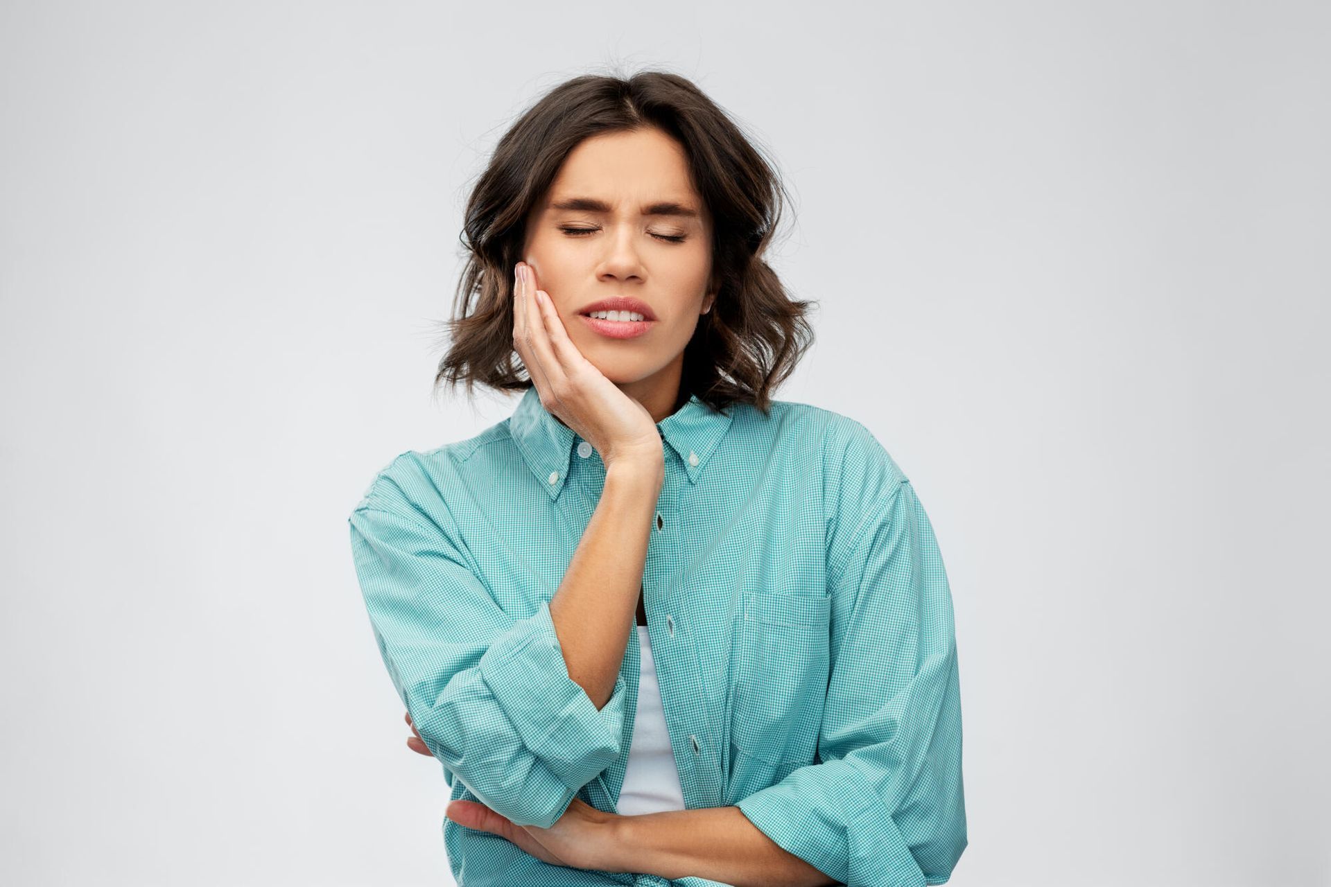 Woman with short hair, eyes closed, holding her jaw in pain. Wearing a turquoise checkered shirt.