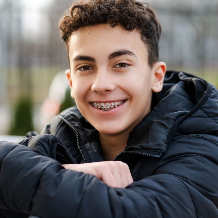 A young man wearing braces and a black jacket smiles for the camera