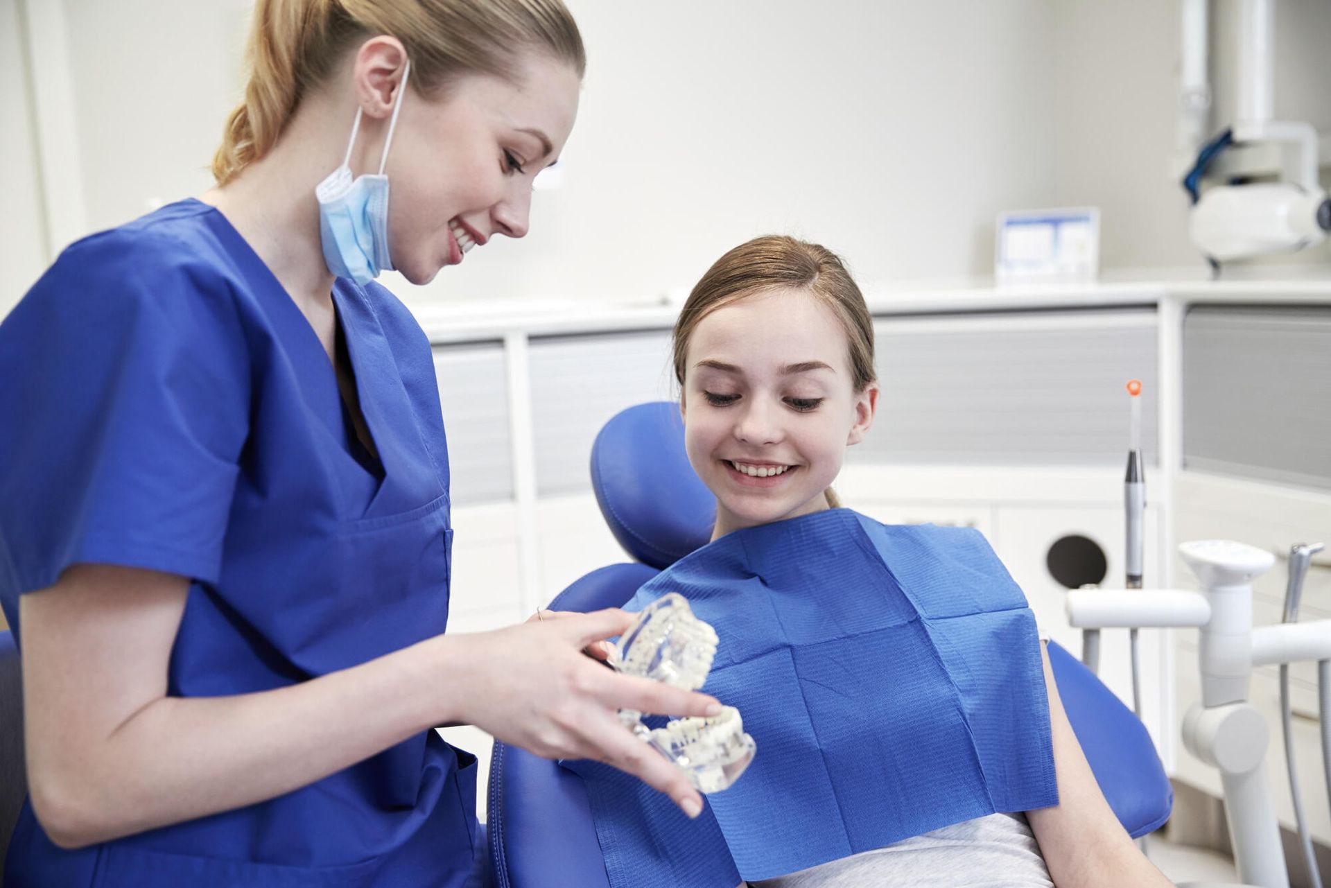 A dental professional in blue scrubs explains a dental model to a patient in a chair.