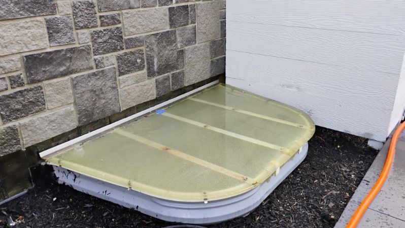 A window well with a cover on it is next to a brick wall.