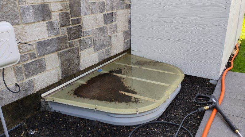 A hose is connected to a window well next to a brick wall.