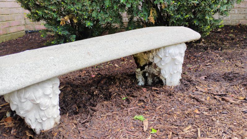 A stone bench is sitting in the dirt in front of a bush.