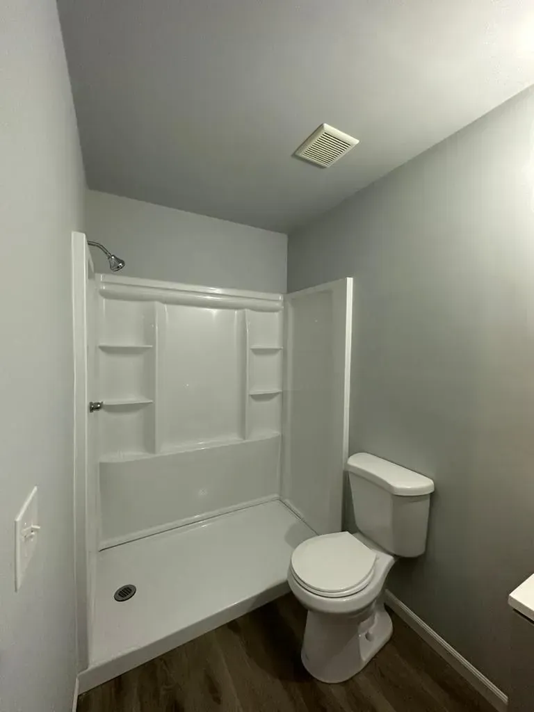 A small, gray-walled bathroom with a white shower stall, toilet, and a dark wood-look floor.