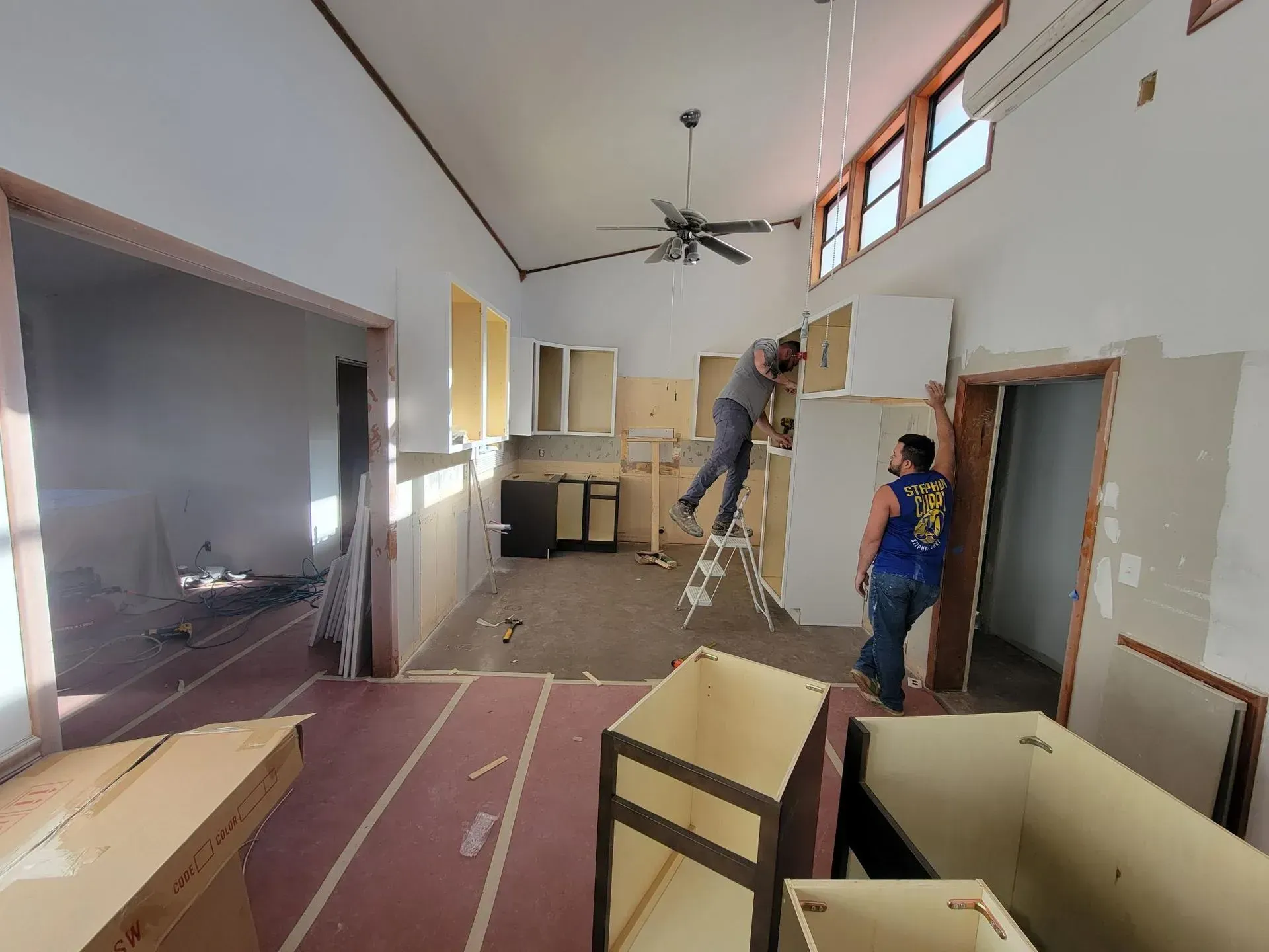 Two people installing cabinets in a room during renovation. Unfinished walls and cabinets, construction debris.