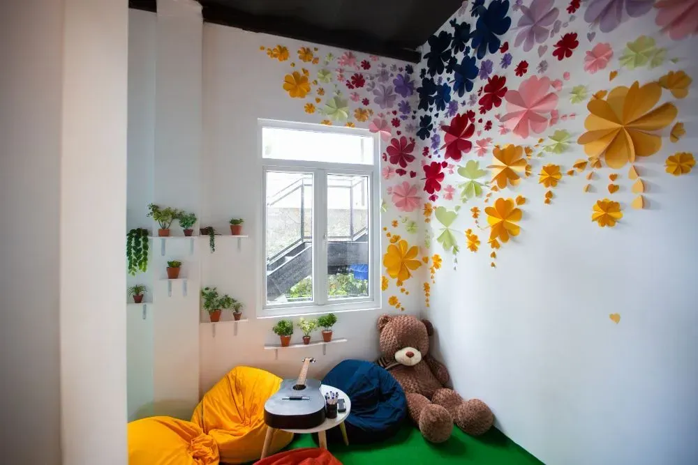 Colorful room with paper flowers on the wall, a window, bean bags, a teddy bear, and plants.