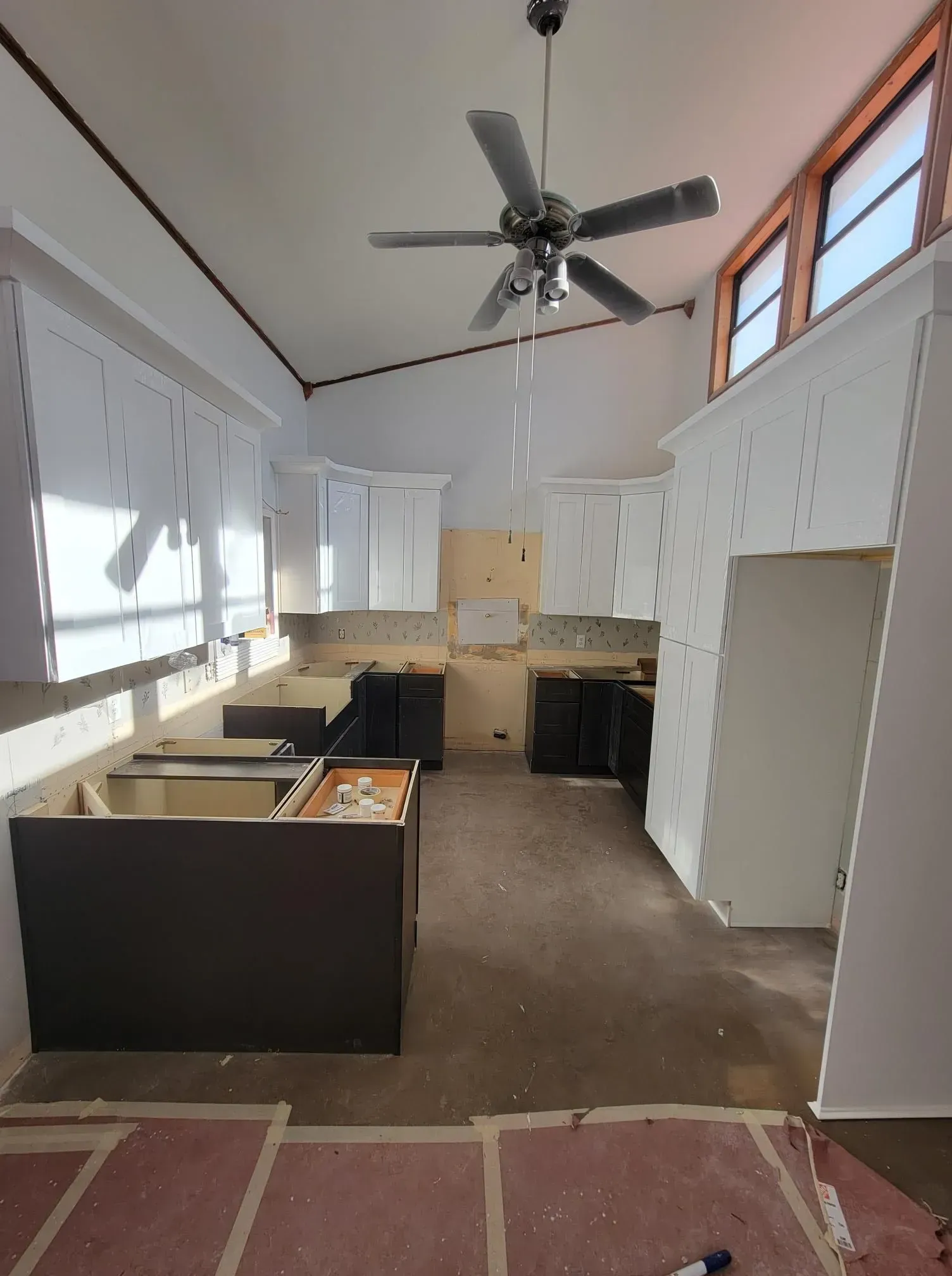 Kitchen under renovation: White cabinets, dark island base, exposed walls, ceiling fan.
