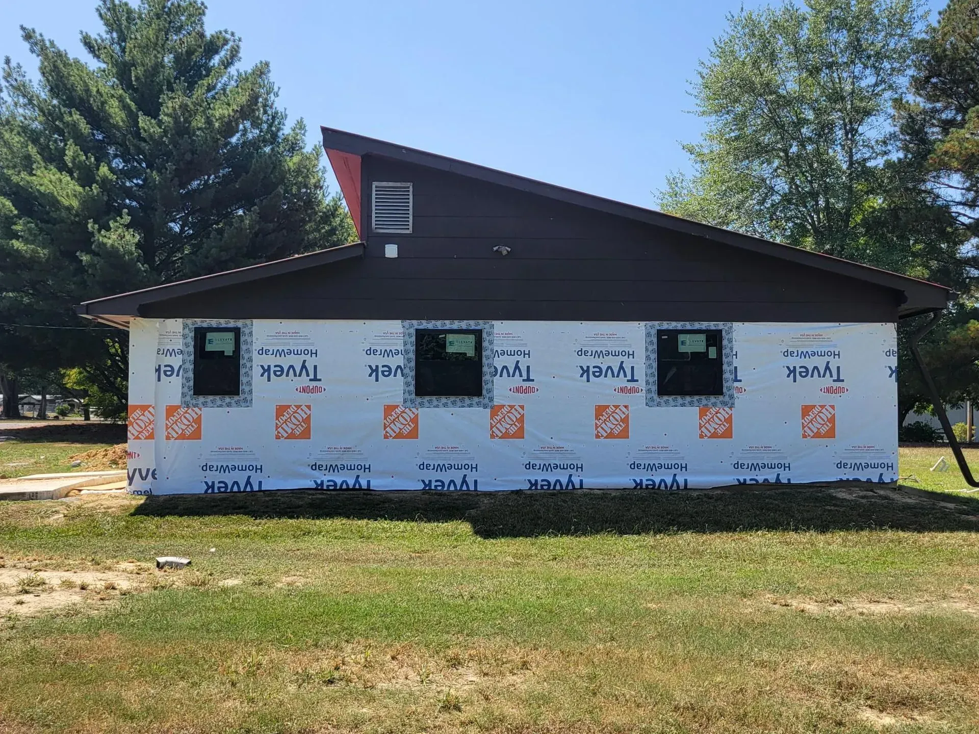 Exterior of a brown-roofed building, windows installed, wrapped in Tyvek, on a grassy lot.