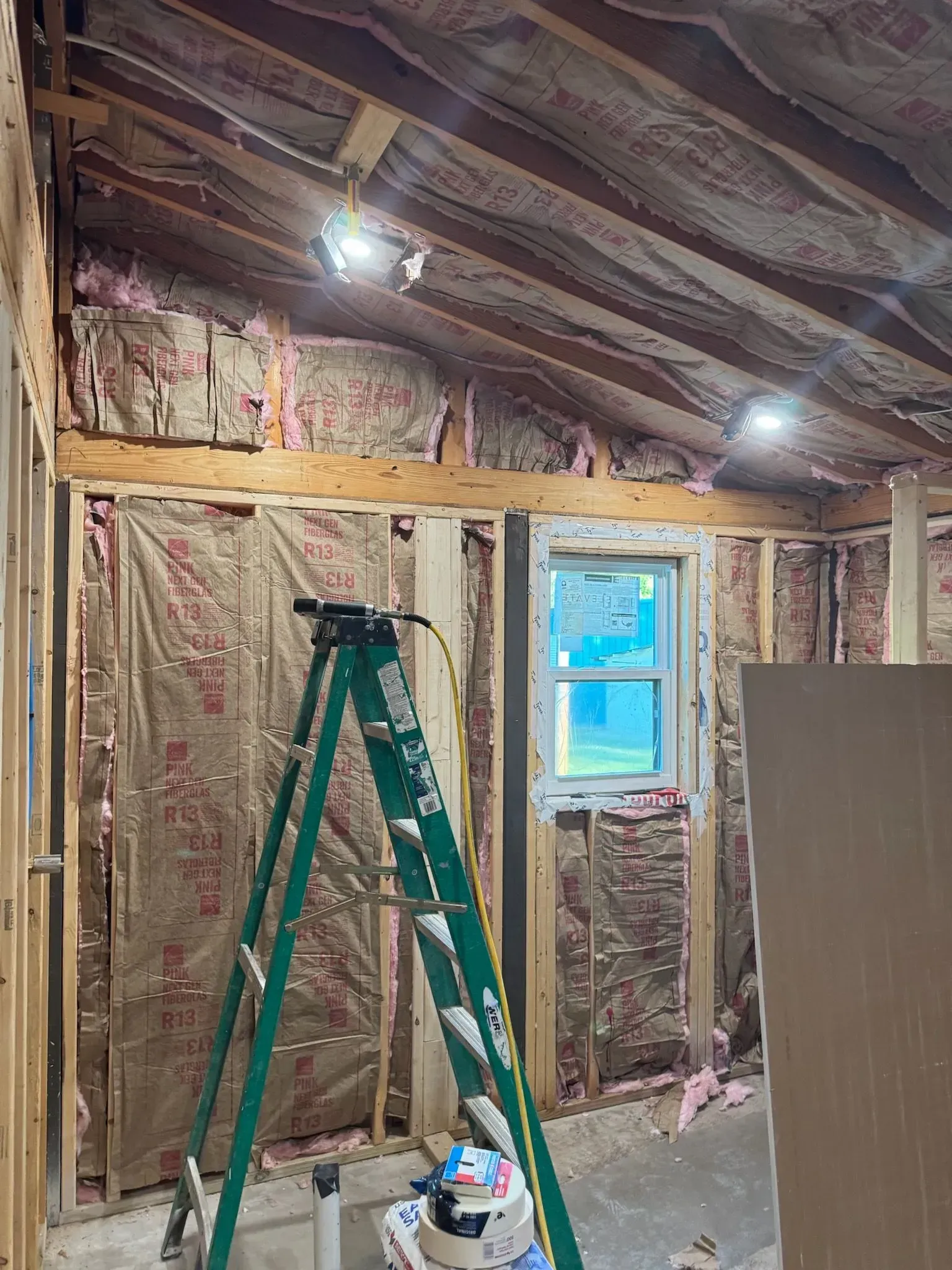 Interior construction view with insulation, studs, and a window. A green ladder is present.