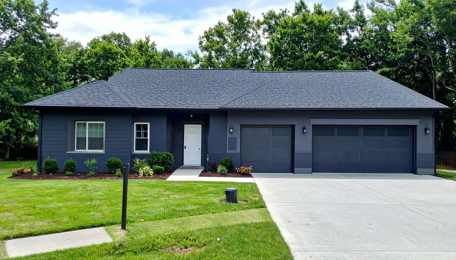 Blue house with black garage doors and roof, light concrete driveway, green lawn and trees.