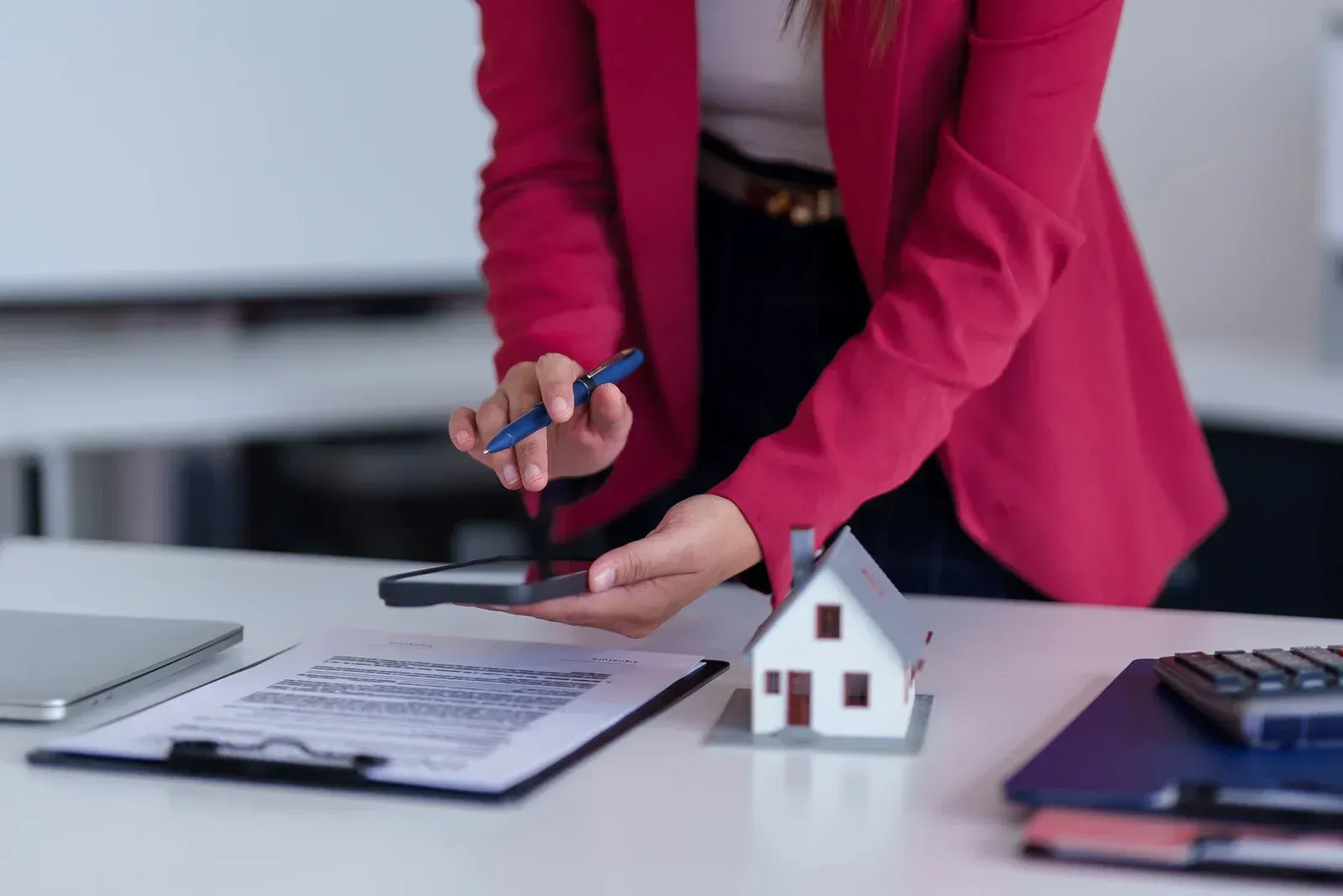 Person in pink blazer uses phone over paperwork and a small house model on a desk.