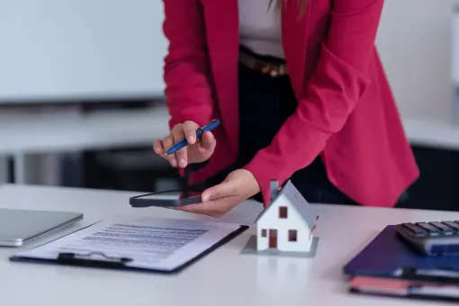 Person in pink blazer uses phone over paperwork and a small house model on a desk.