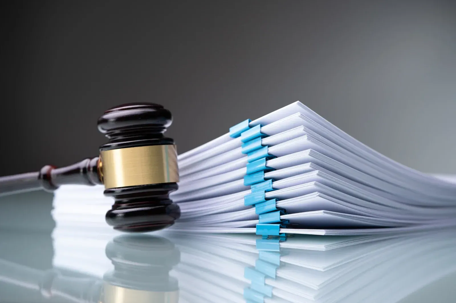 Gavel next to a stack of legal documents, held together by blue binder clips, on a reflective surface.