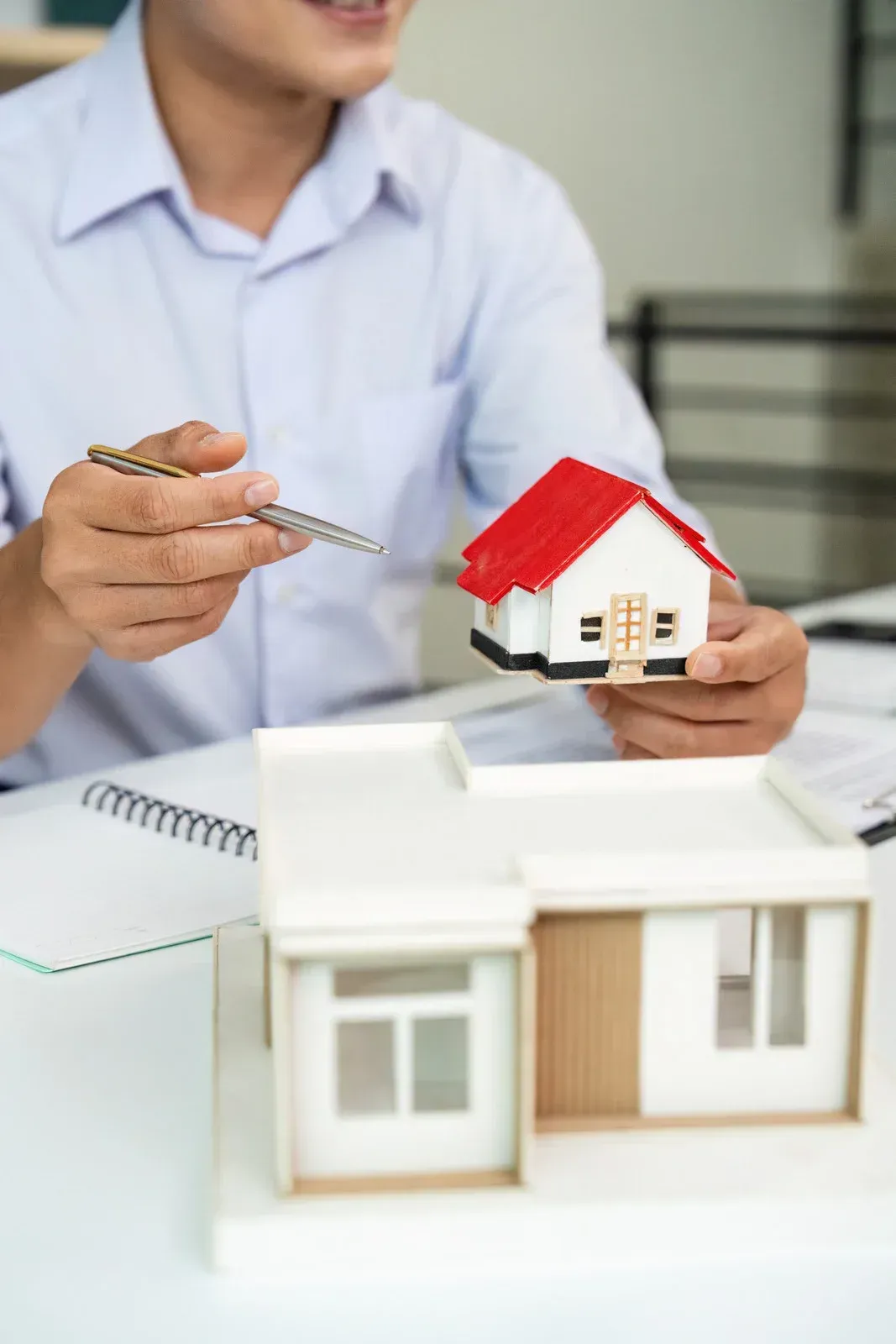 Man holding a small house model, pointing with a pen, near a larger house model.
