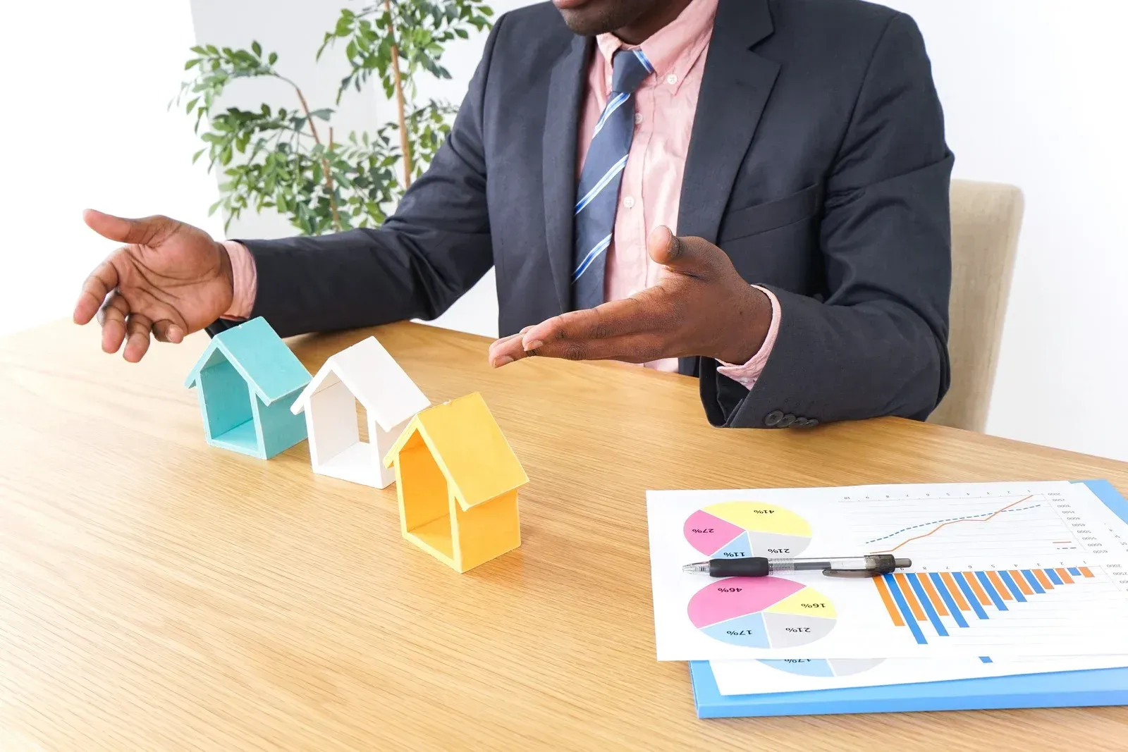 A man in a suit gestures toward three miniature houses on a table with charts, possibly real estate options.