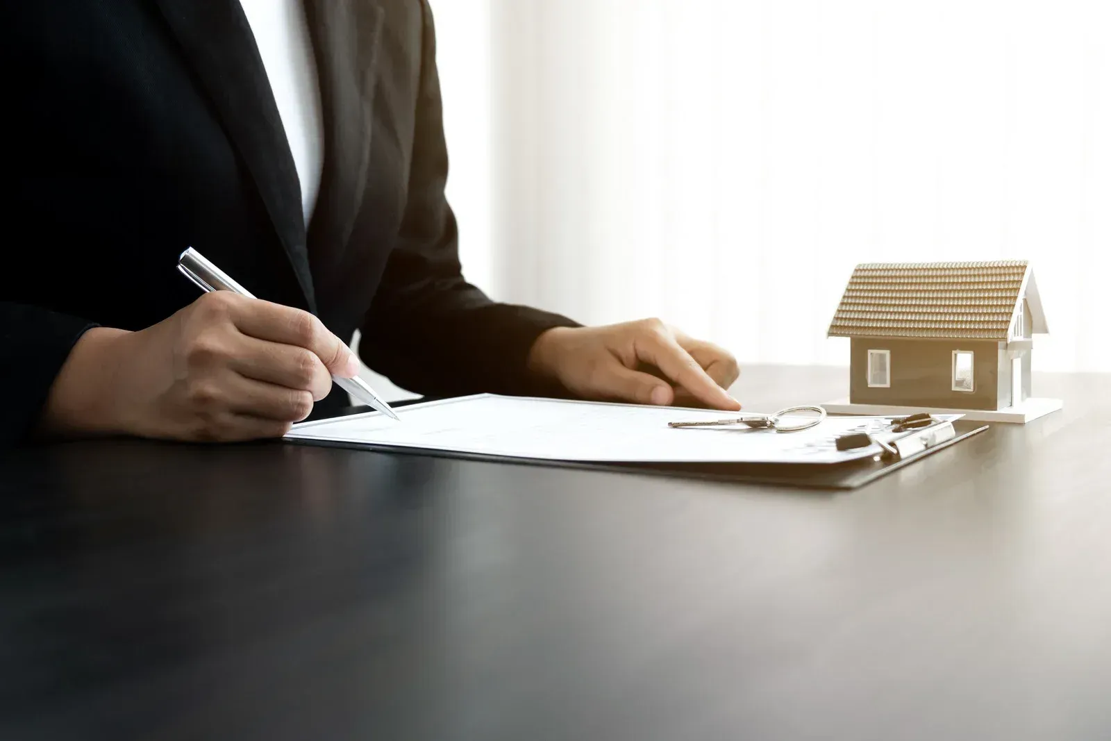 Person in a blazer signing paperwork with a small house model on a table.