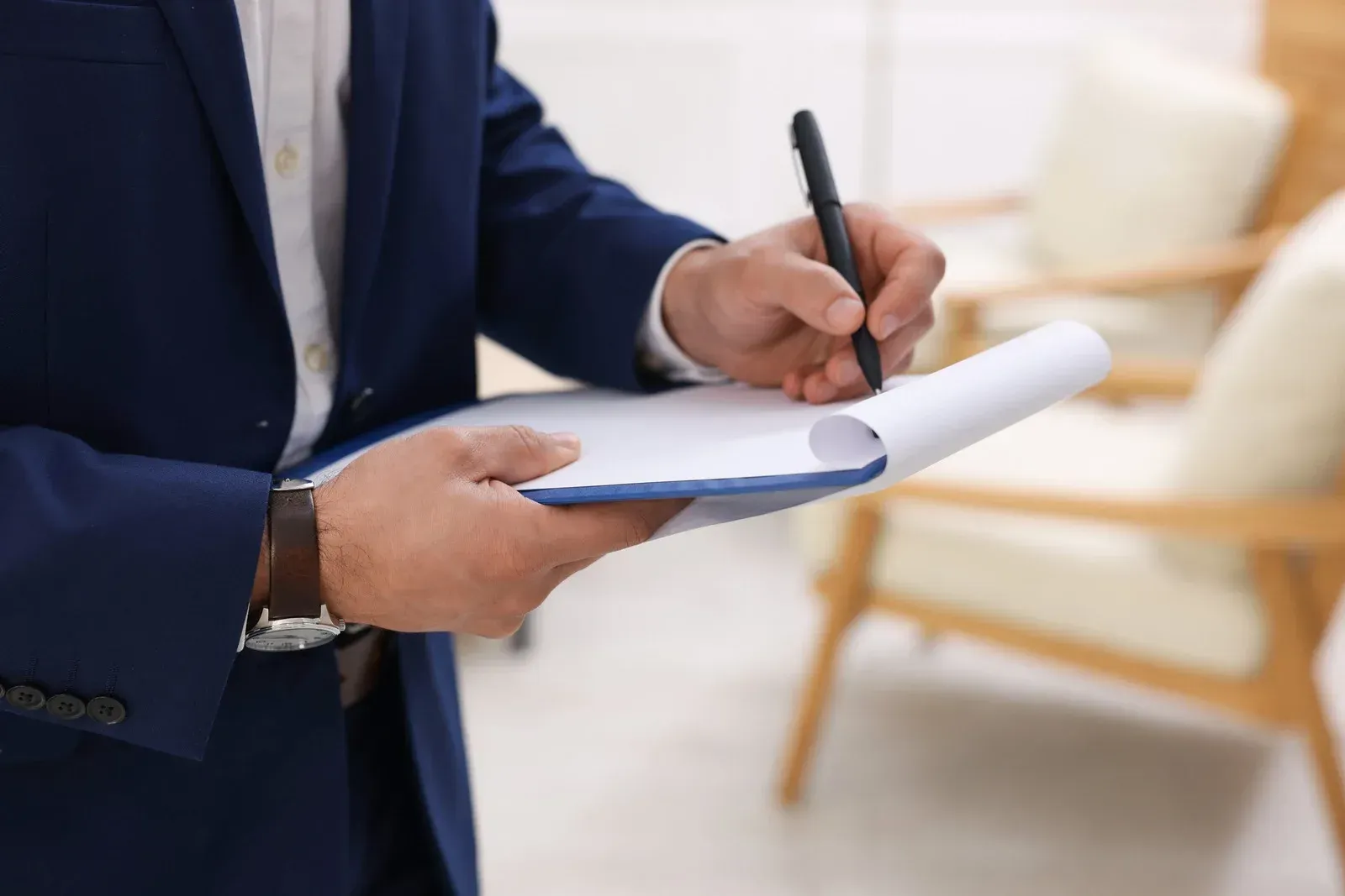 Person in blue suit writing on clipboard, indoors with chairs in background.