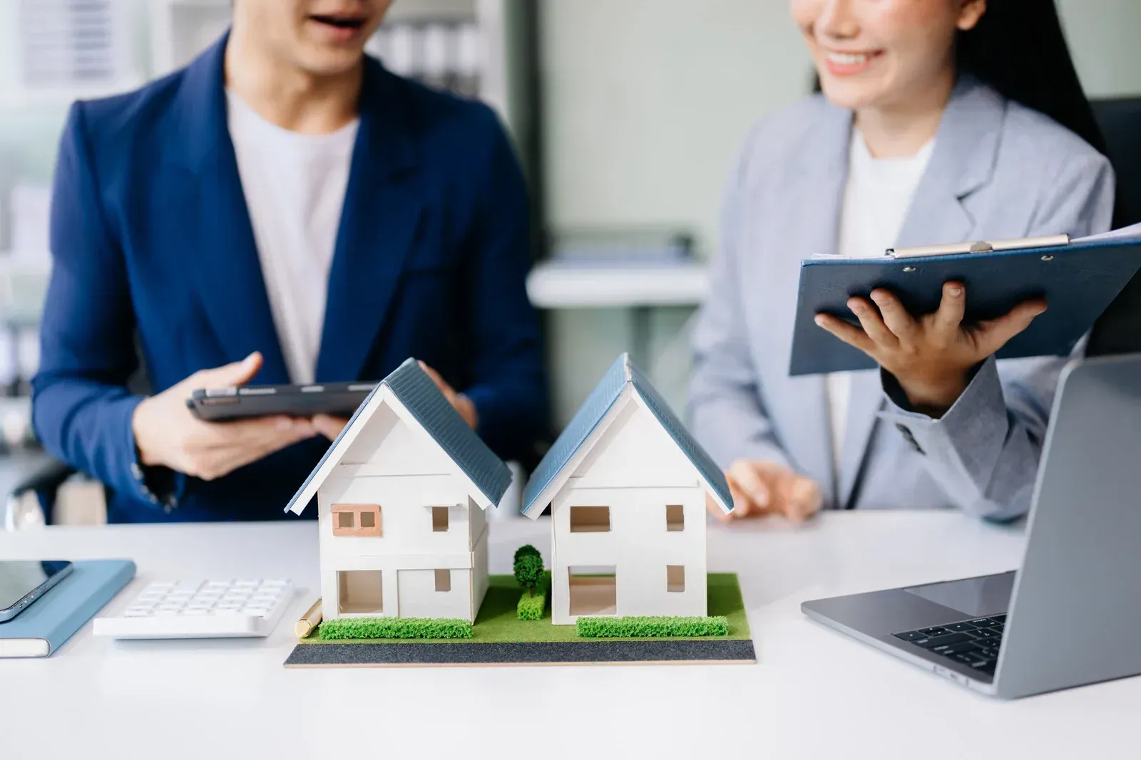 Two people discussing housing models; desk with laptop, tablet, and calculator.