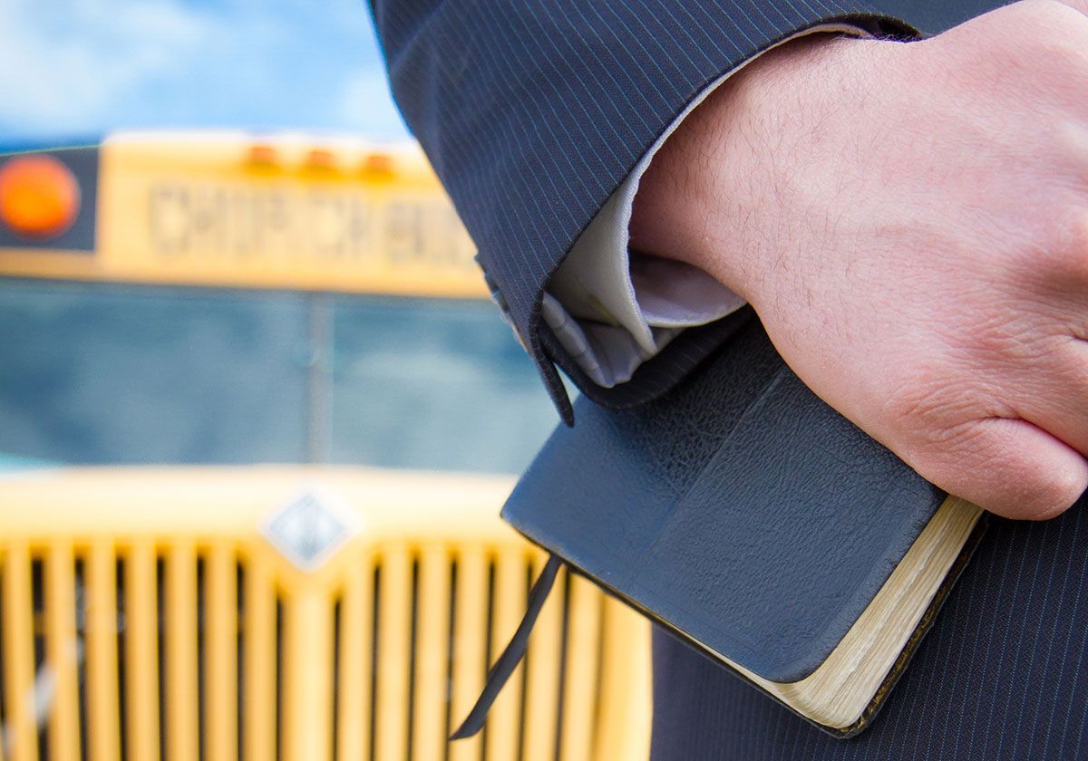 A close-up view of a person in a suit holding a small, dark, bound book with a yellow school bus in the background.