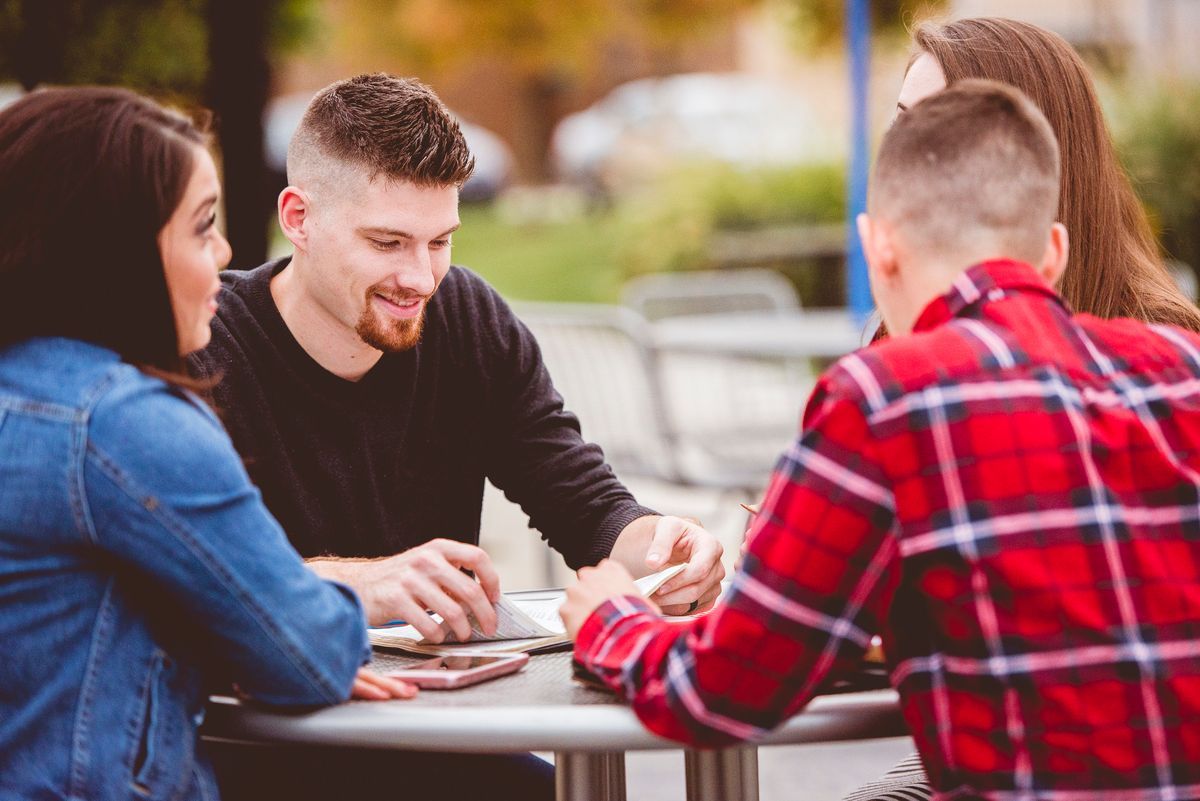 Four people sit at an outdoor table, smiling and working together on papers.