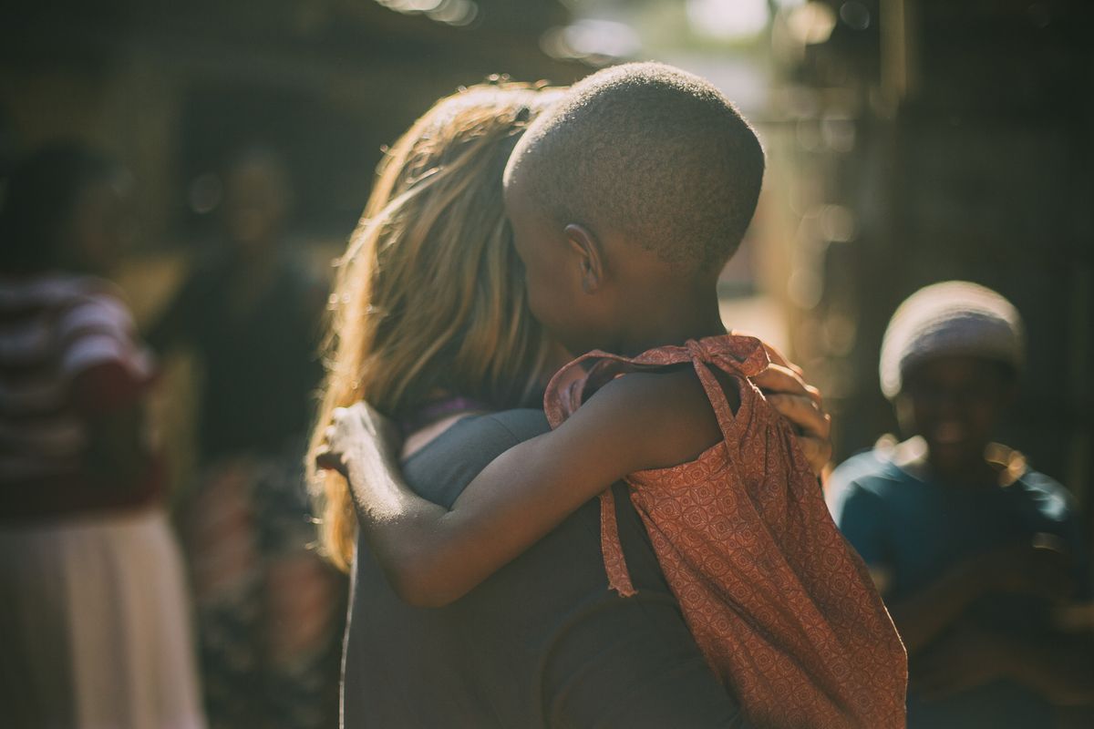 A person in a gray shirt embraces a child wearing a red top, with blurred figures in a warm-toned outdoor setting.