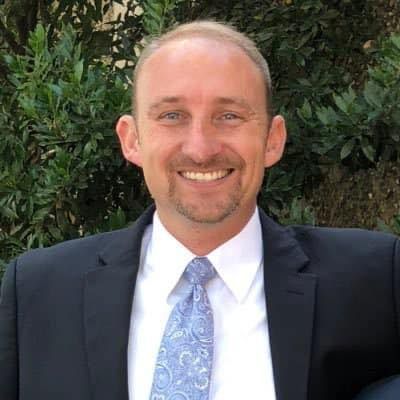 A person in a dark suit, white collared shirt, and light blue patterned tie smiles in front of green foliage.