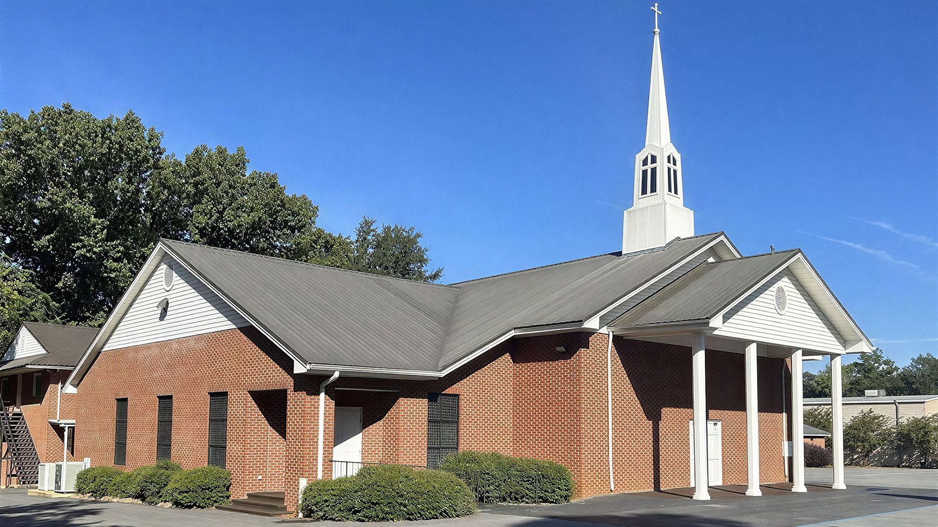 A red brick church building with a white steeple, covered porch, and columns under a clear blue sky.