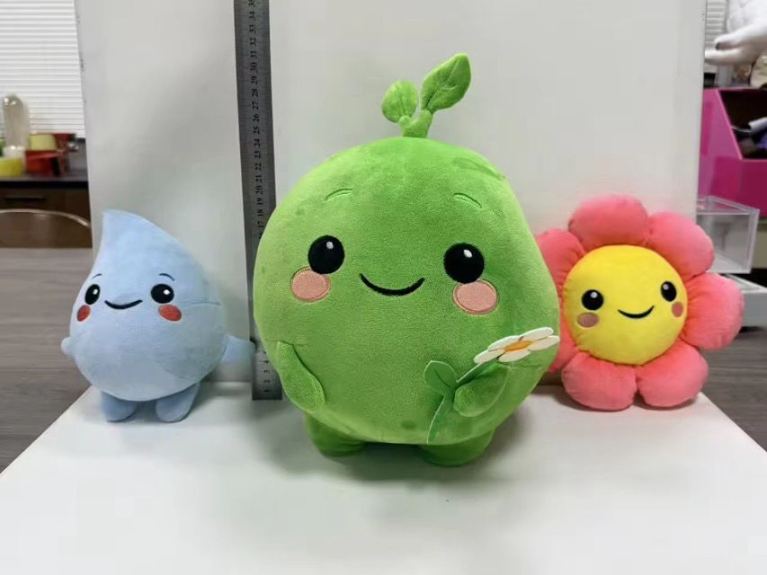 Three plush toys: a blue water drop, a large green round creature holding a flower, and a pink and yellow flower.