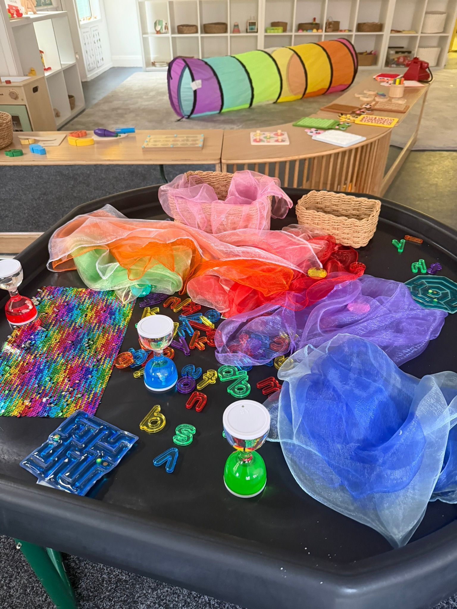 A tray filled with colorful sensory scarves, transparent gems, and small containers, located in a bright playroom.