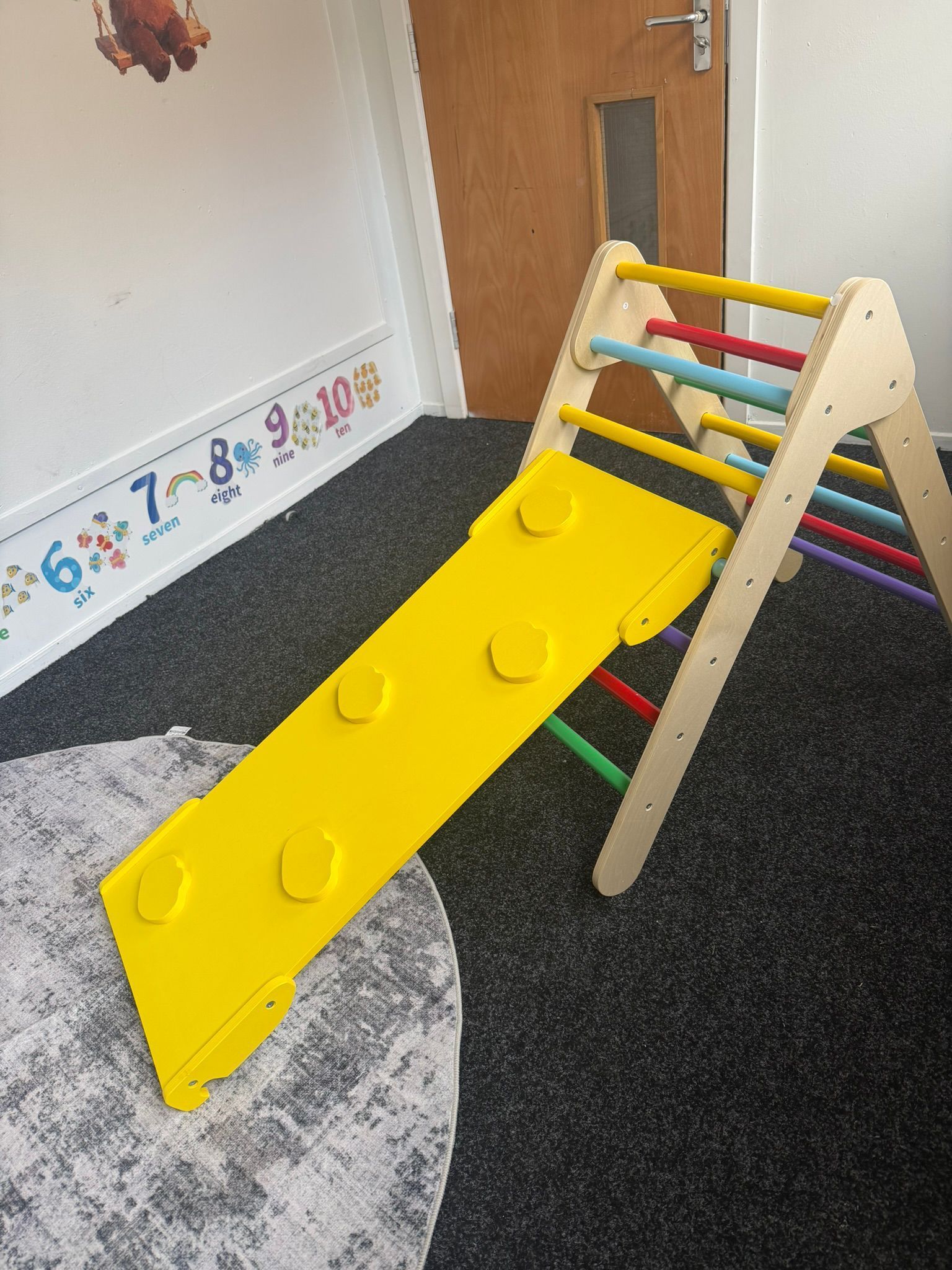 A wooden Pikler triangle climbing frame with a yellow slide attached, positioned on a gray rug in a room.
