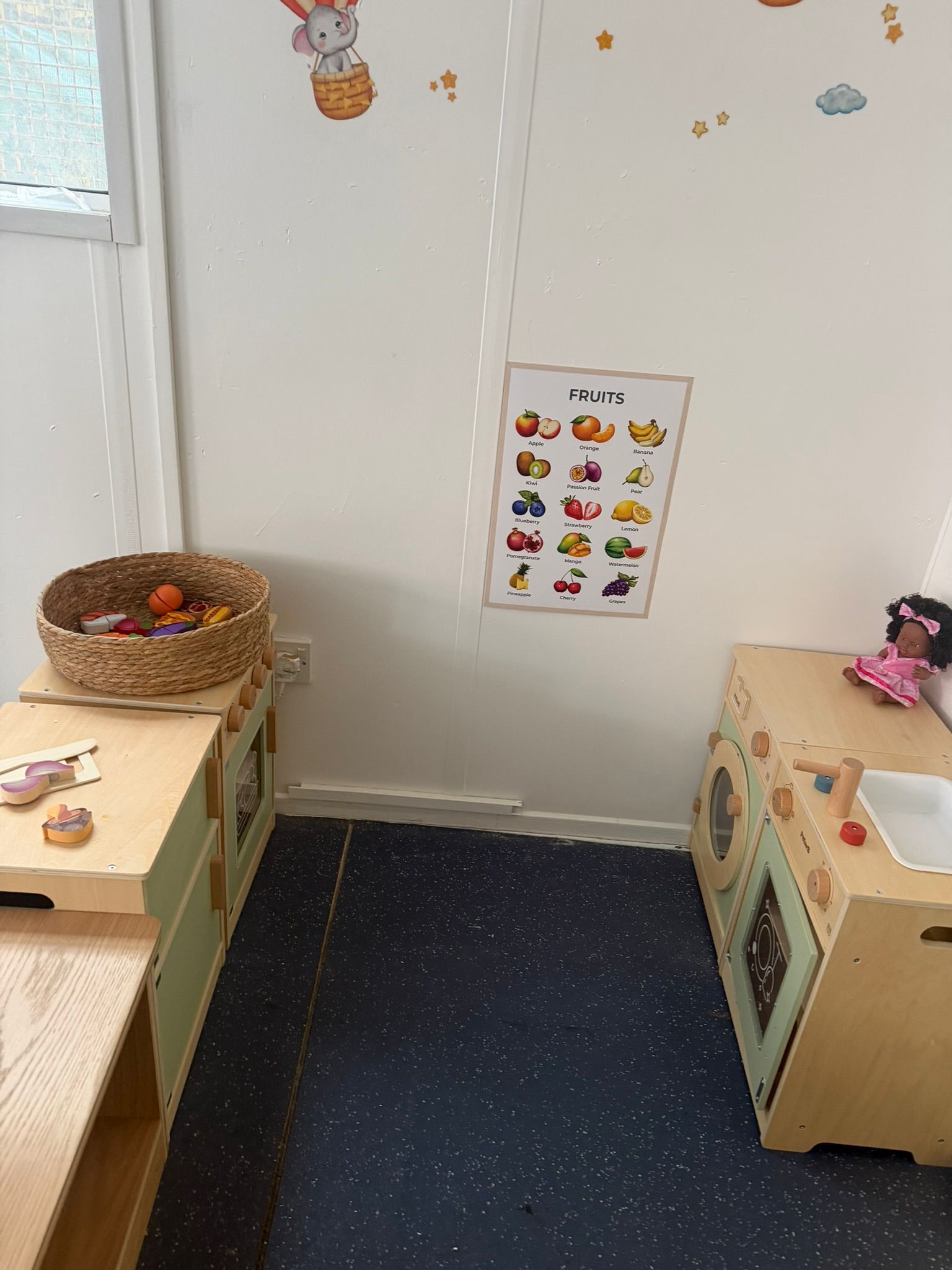 A child's playroom corner with two wooden play stations, a woven basket, a wall poster of animals, and a doll on the right.