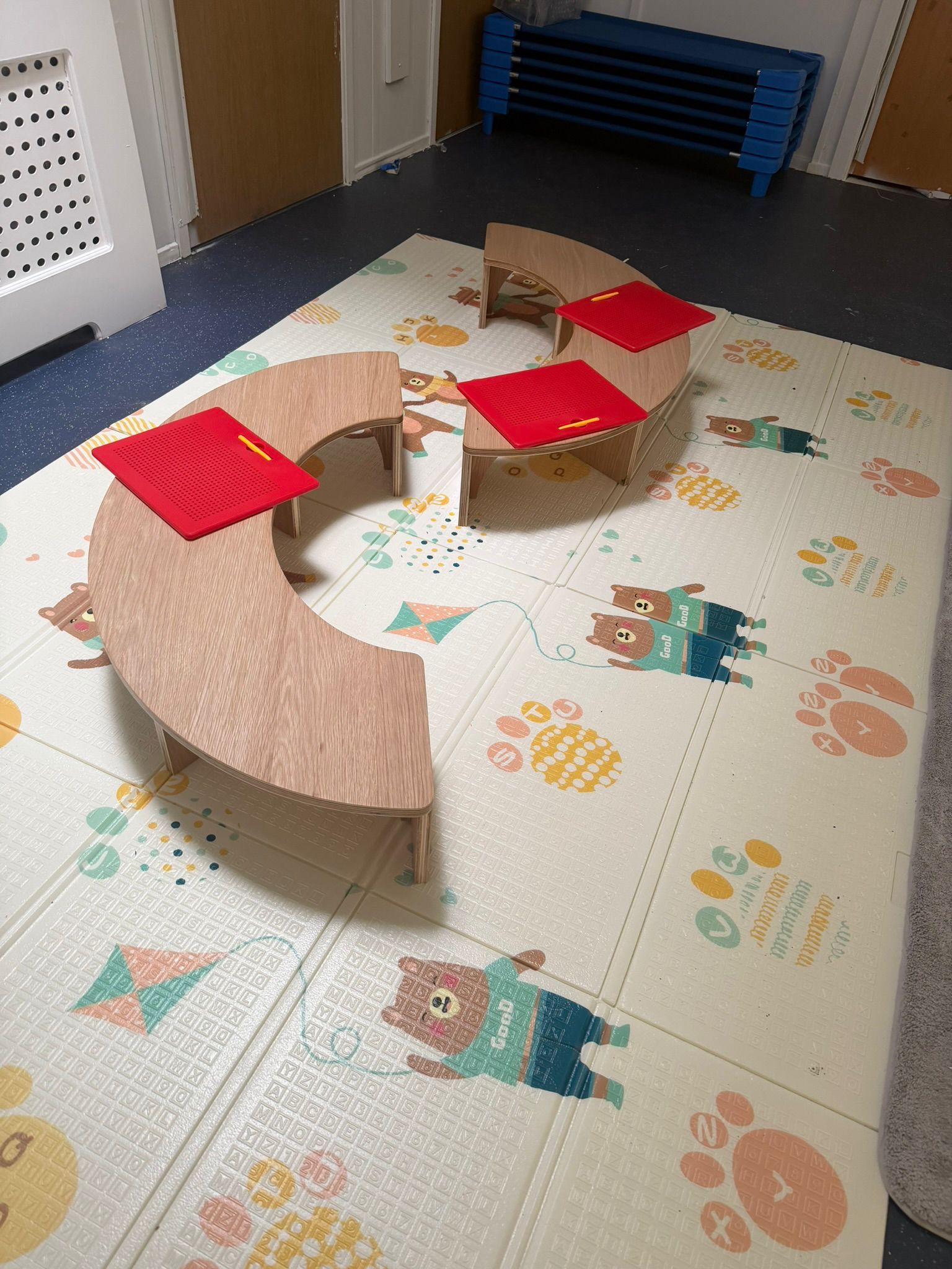 Curved wooden children's tables with red mats on a floor mat decorated with cartoon bear illustrations.
