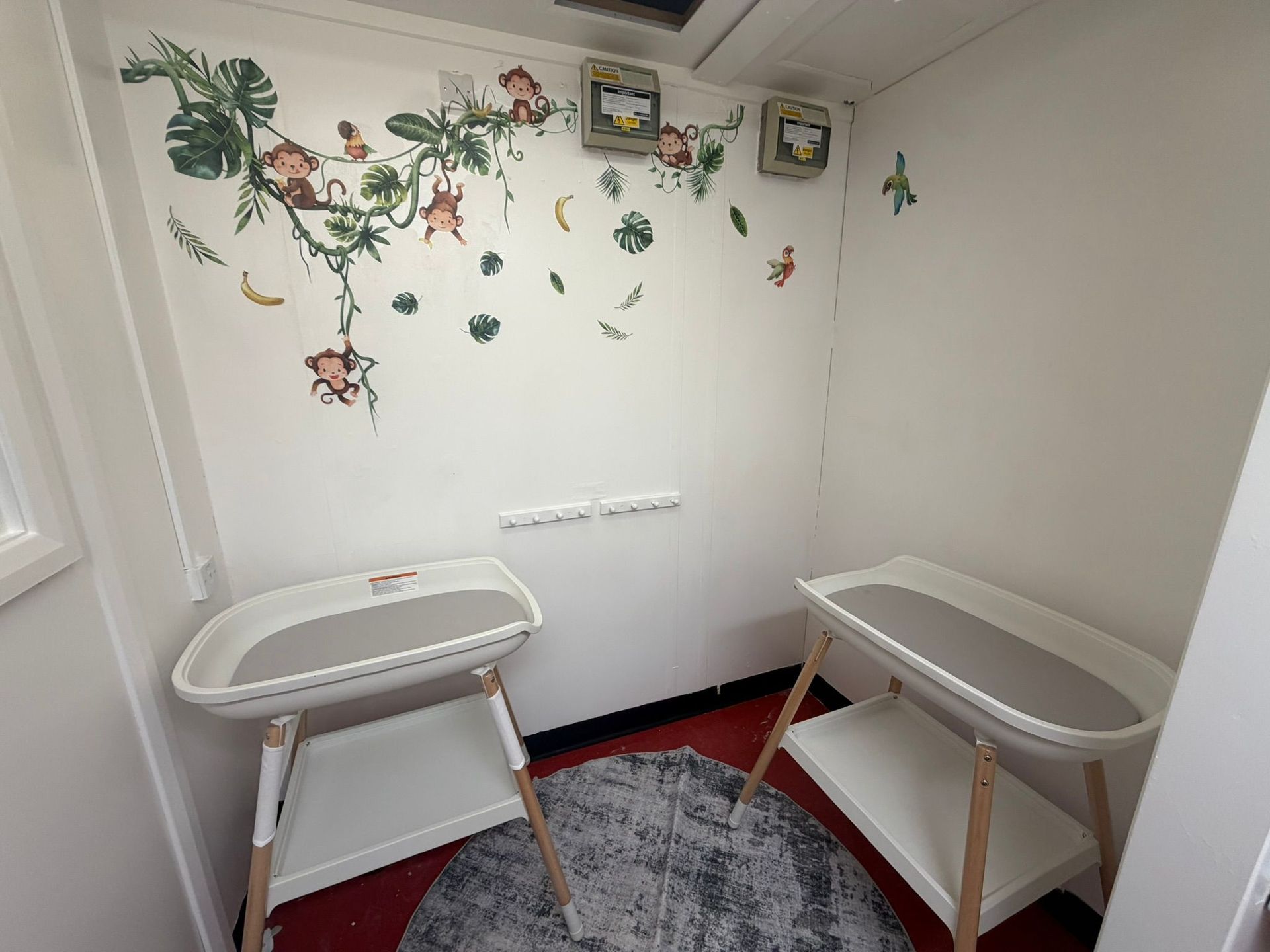Two white changing tables stand on a grey rug in a corner decorated with a jungle-themed monkey wall decal.