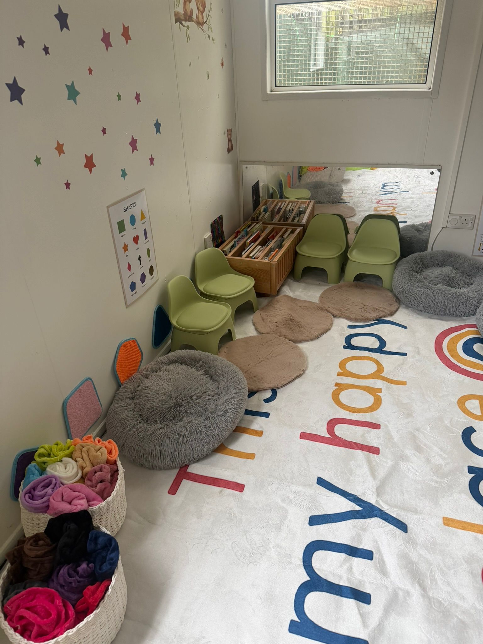 A cozy corner for children featuring small green chairs, textured seating pads, books, and colorful baskets of fabrics.