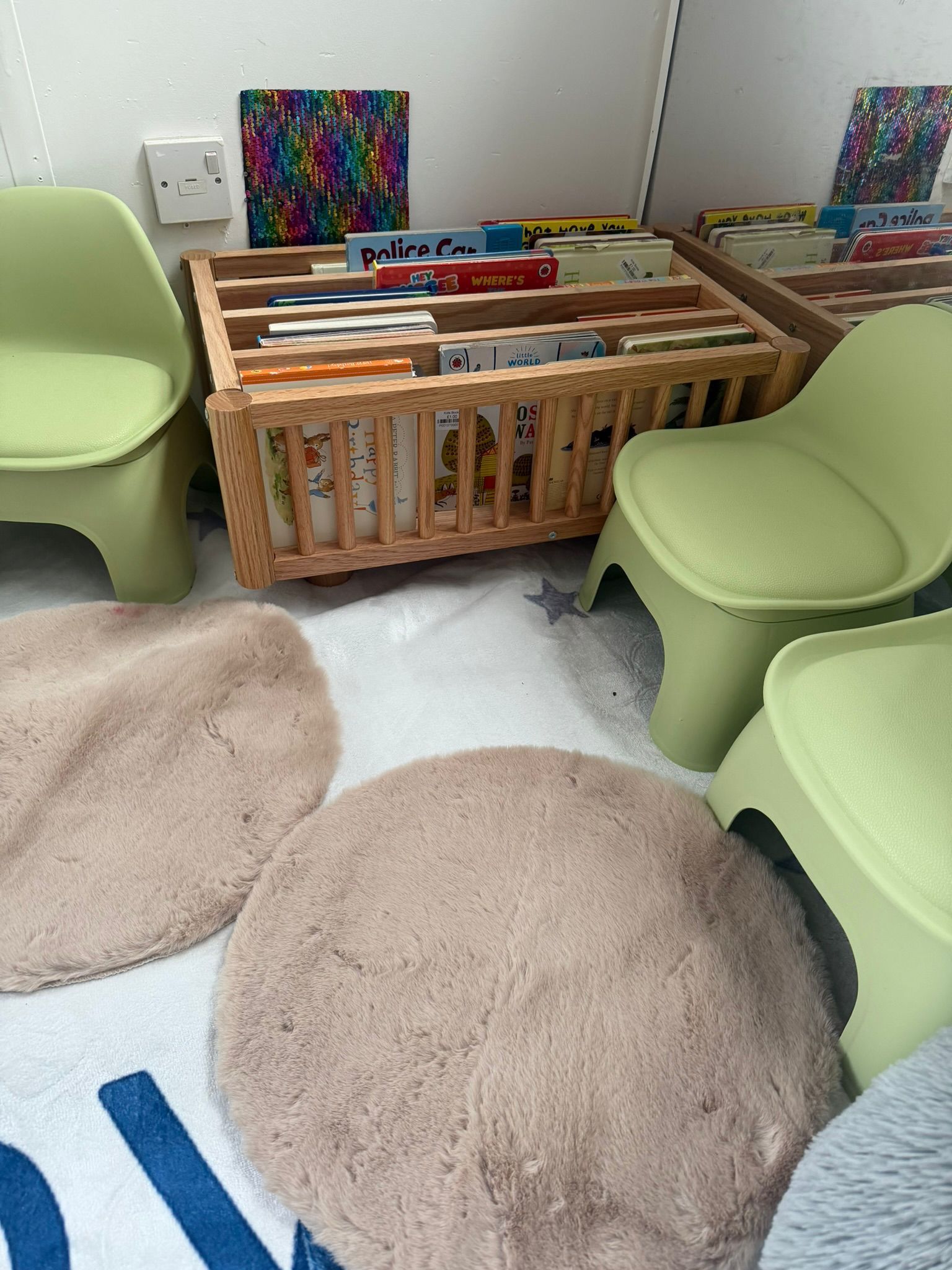 A wooden children’s bookshelf filled with books, surrounded by light green plastic chairs and plush, round floor mats.