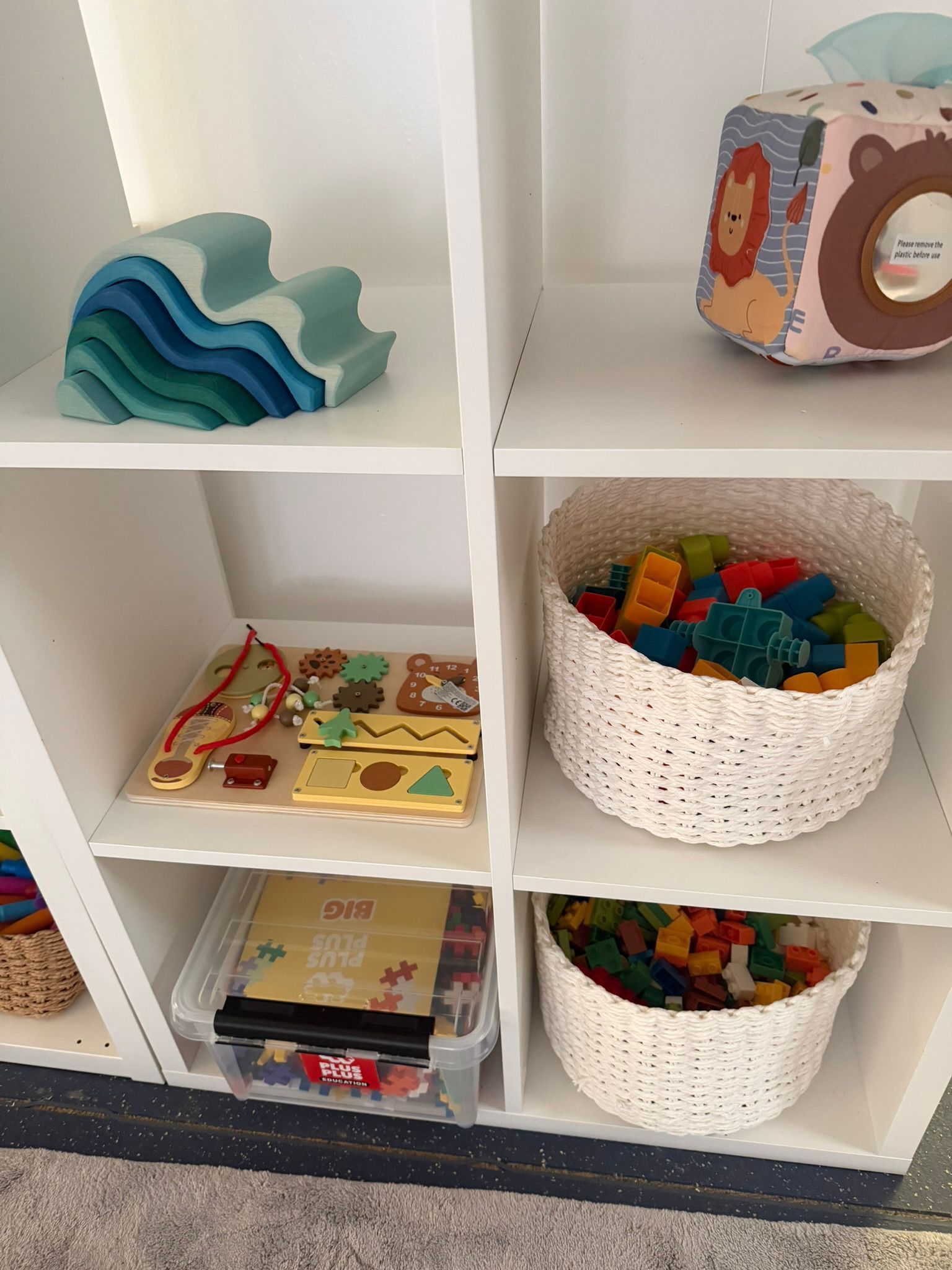 A white, multi-shelf storage unit holding various children's toys, including building blocks in baskets and a wave toy.