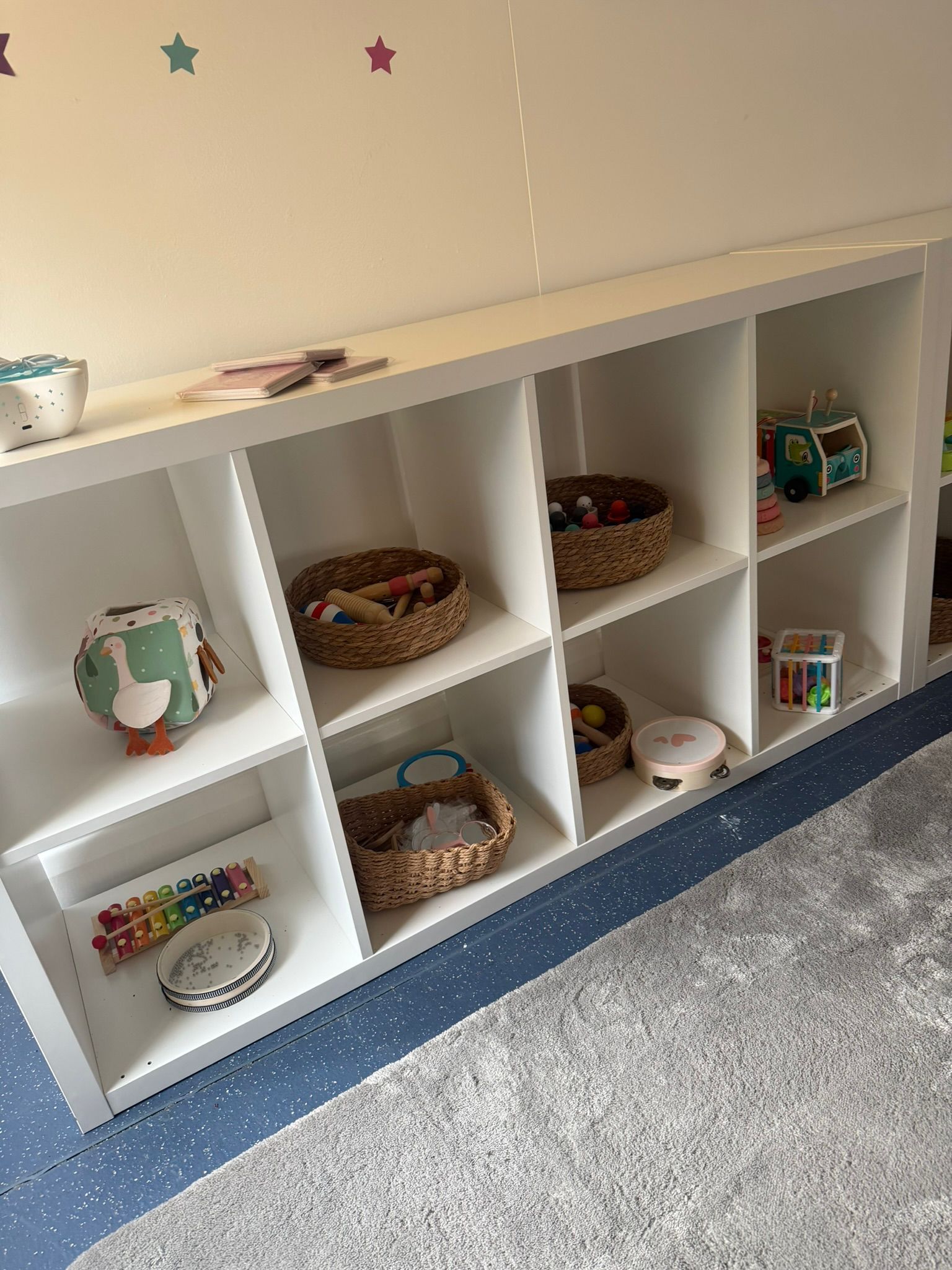 A white cube storage shelf containing various children's toys in woven baskets and on open shelves, sitting on a rug.