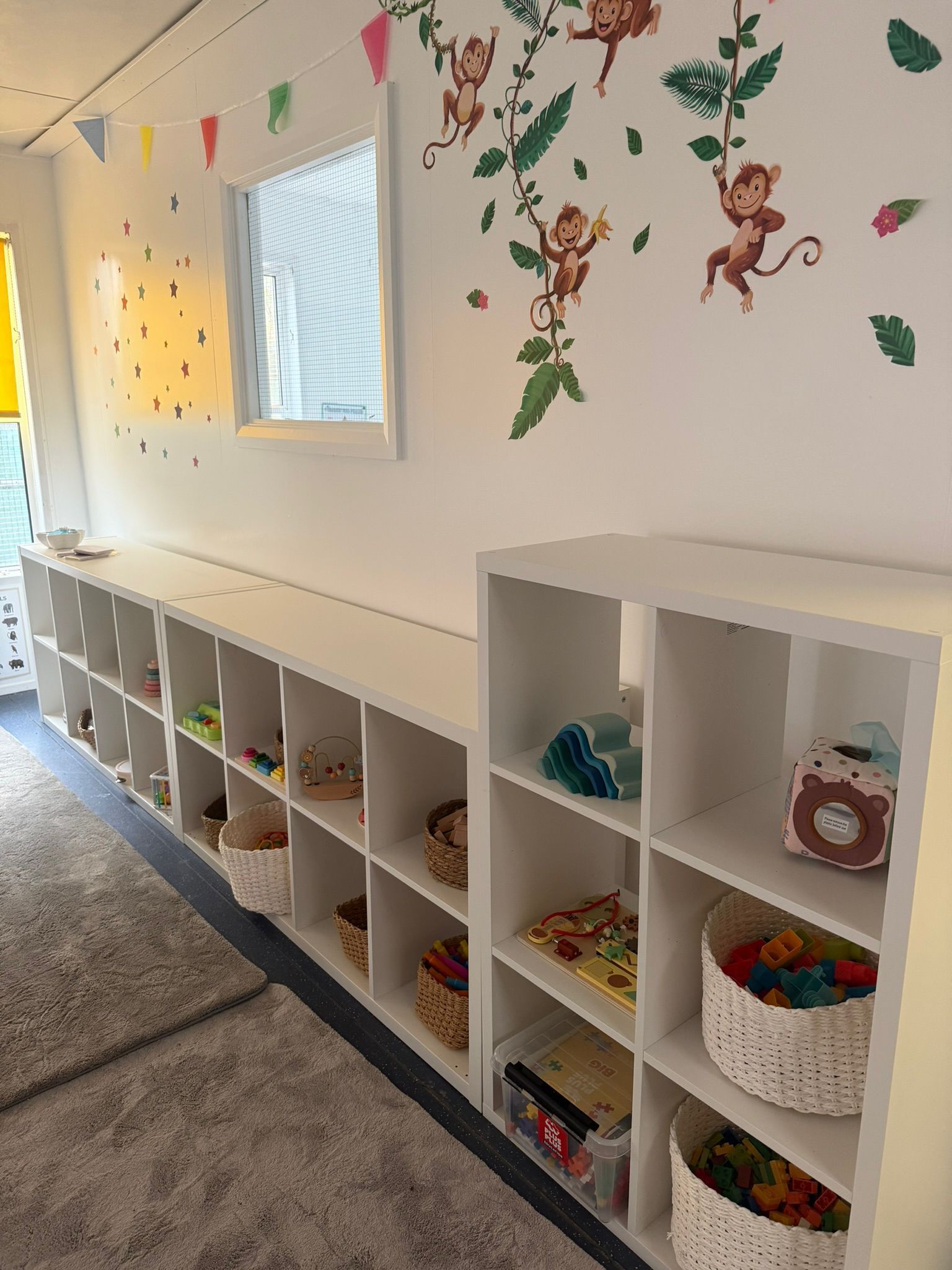 Low white shelving units holding toys in baskets and bins, set against a wall with playful monkey decals.