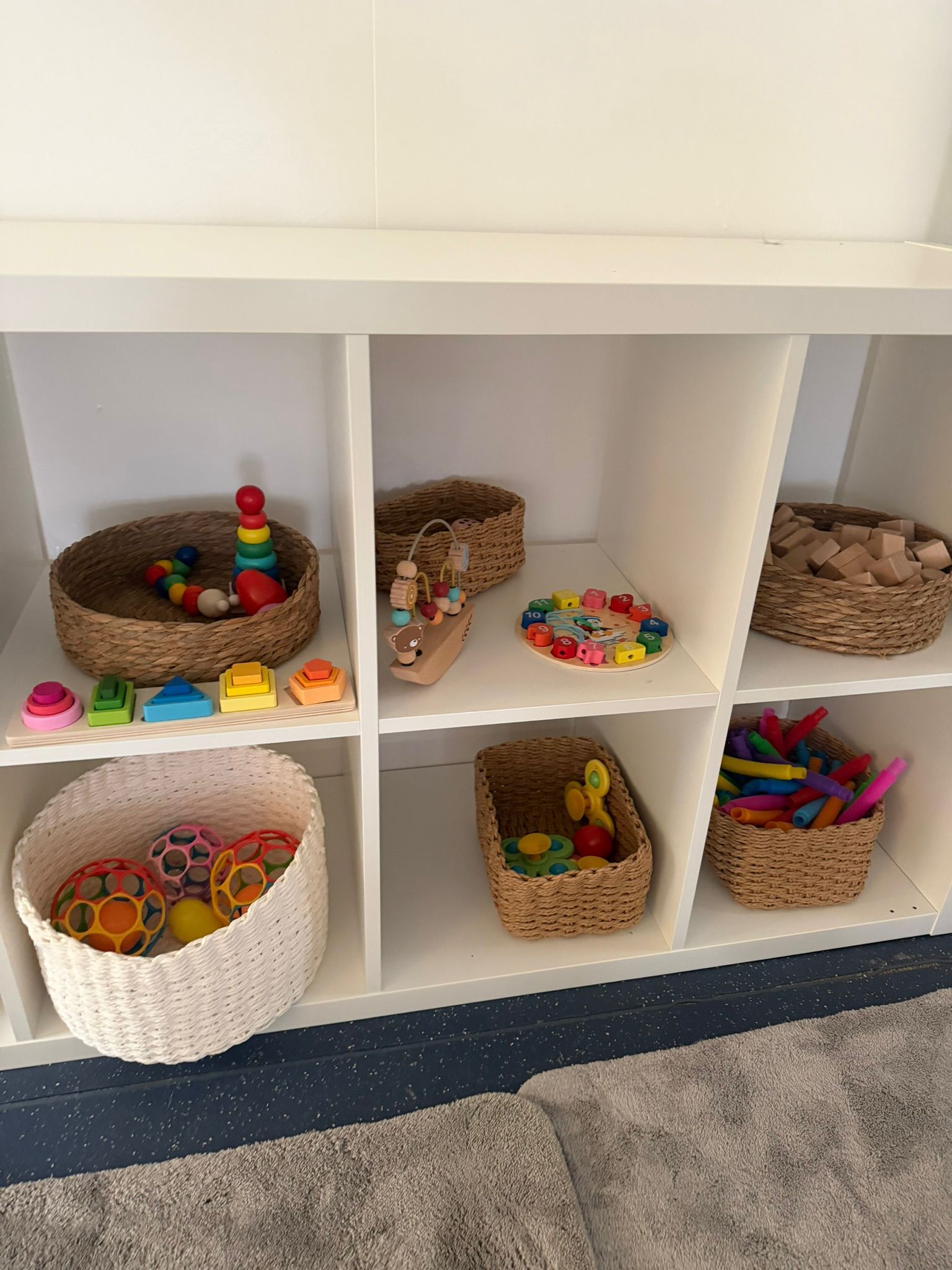 A white cubby storage unit holds several woven baskets and open bins containing colorful children's toys.