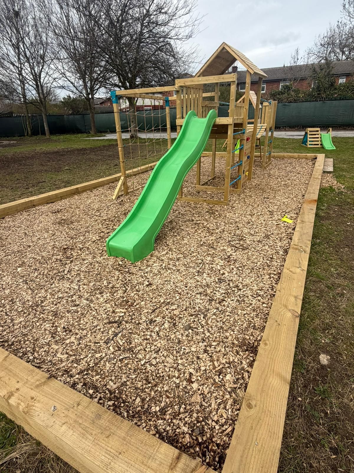 A wooden playground structure with a green slide sits inside a rectangular border filled with wood chips on a grass field.