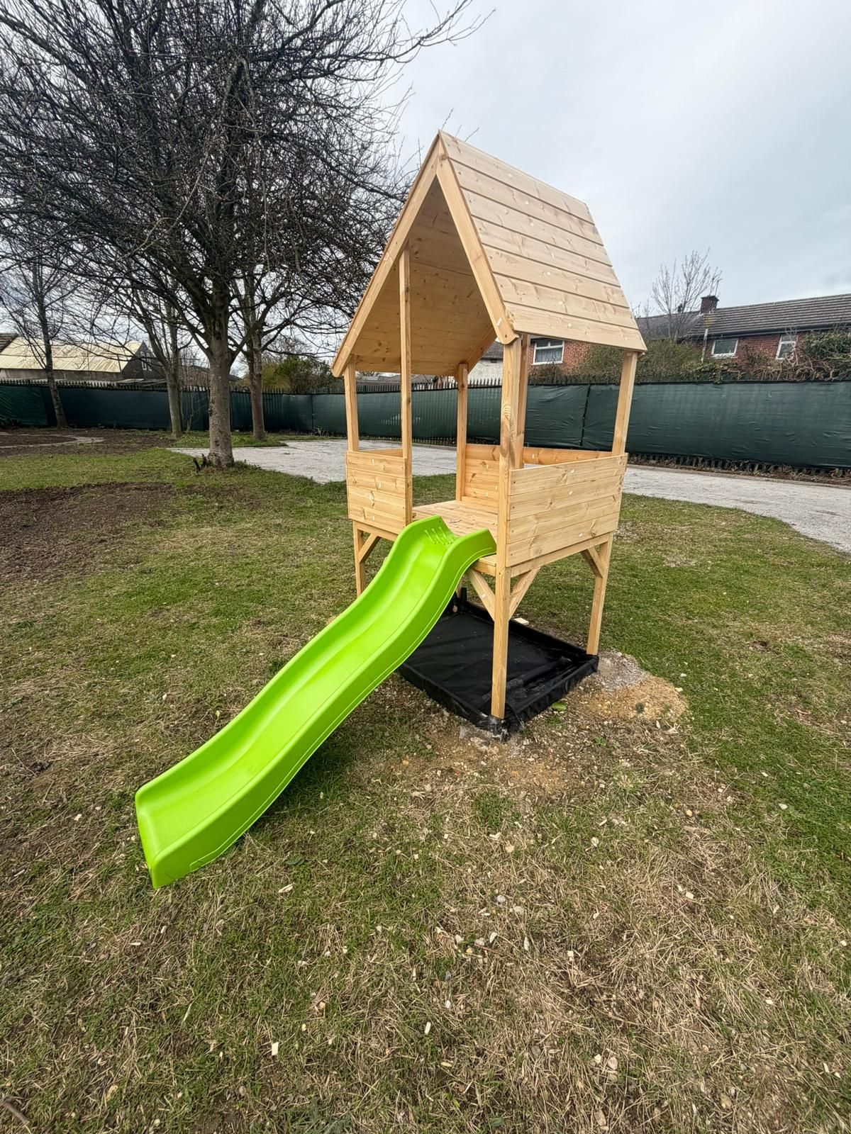 A wooden playhouse on stilts with a bright green slide, situated in a backyard with a grass lawn and a green fence.
