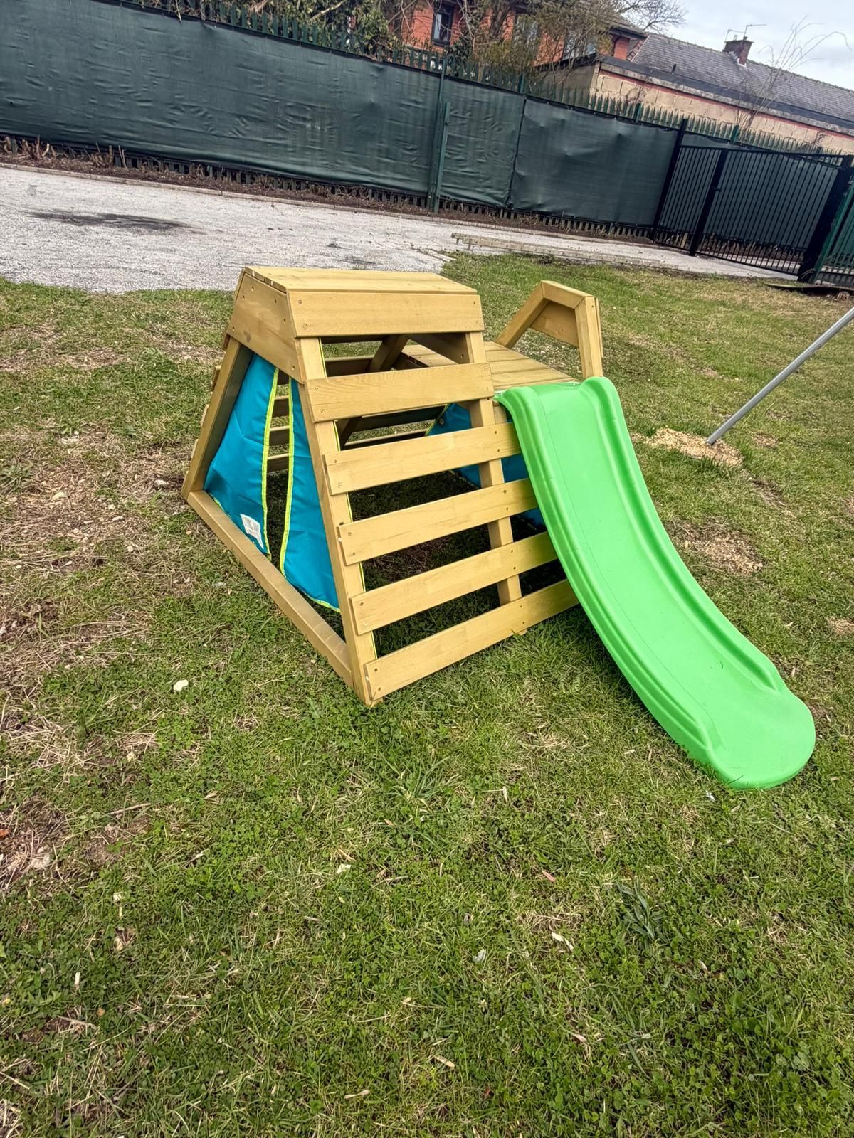 A wooden children’s play structure with a teal fabric tunnel and a lime green slide on a grass lawn.