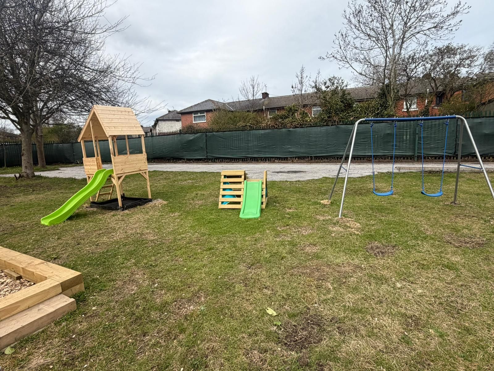 A playground on a grassy area featuring a wooden playhouse with a green slide, a small climbing frame, and a swing set.