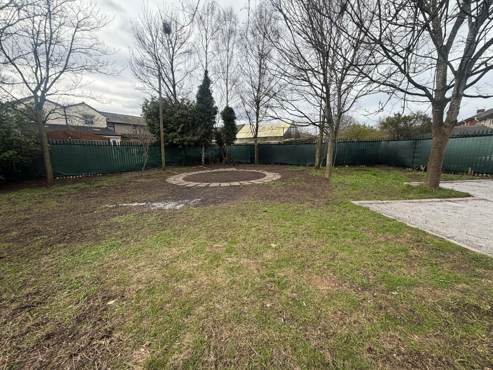 A large backyard with patches of grass and dirt, a circular paved feature, bare trees, and a green mesh fence.