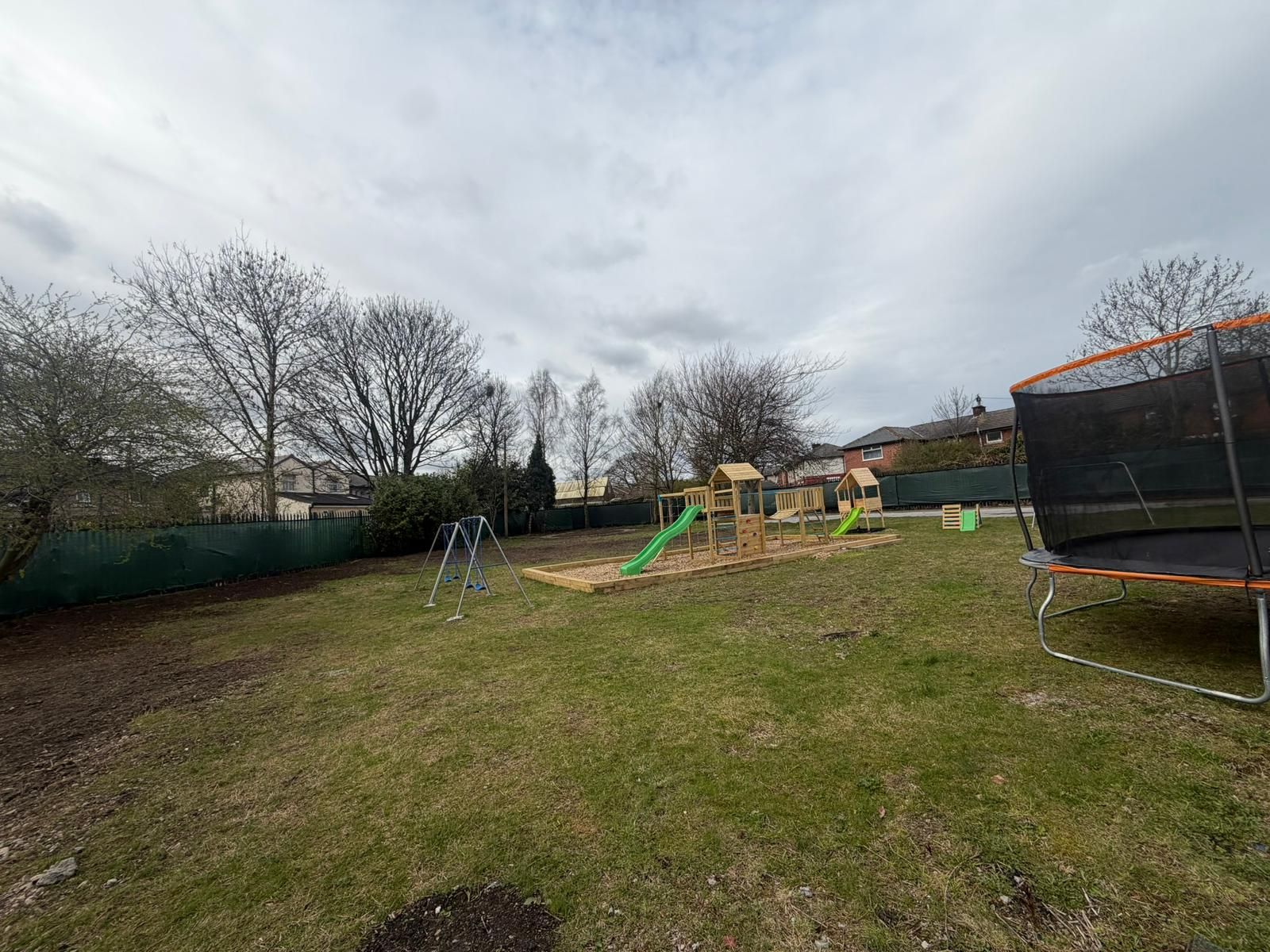 A backyard with a wooden play structure featuring two slides, a small trampoline on the right, and trees under a cloudy sky.