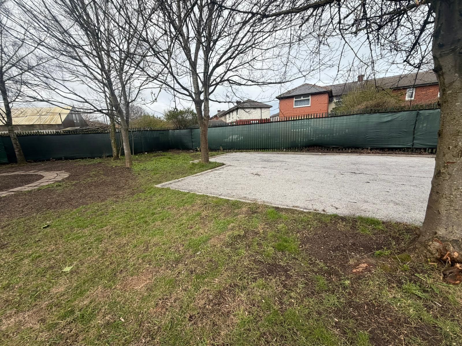A grassy backyard featuring a patch of gravel, leafless trees, and a green privacy fence in front of distant houses.