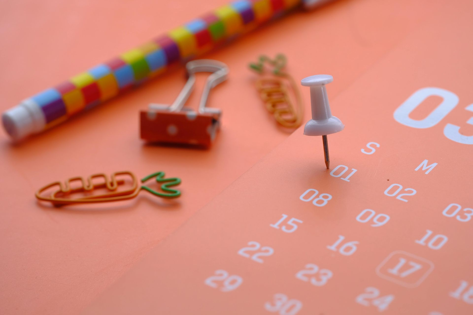 A thumbtack pinned on the 1st of a coral-colored calendar, next to a colorful patterned pen and two carrot-shaped clips.