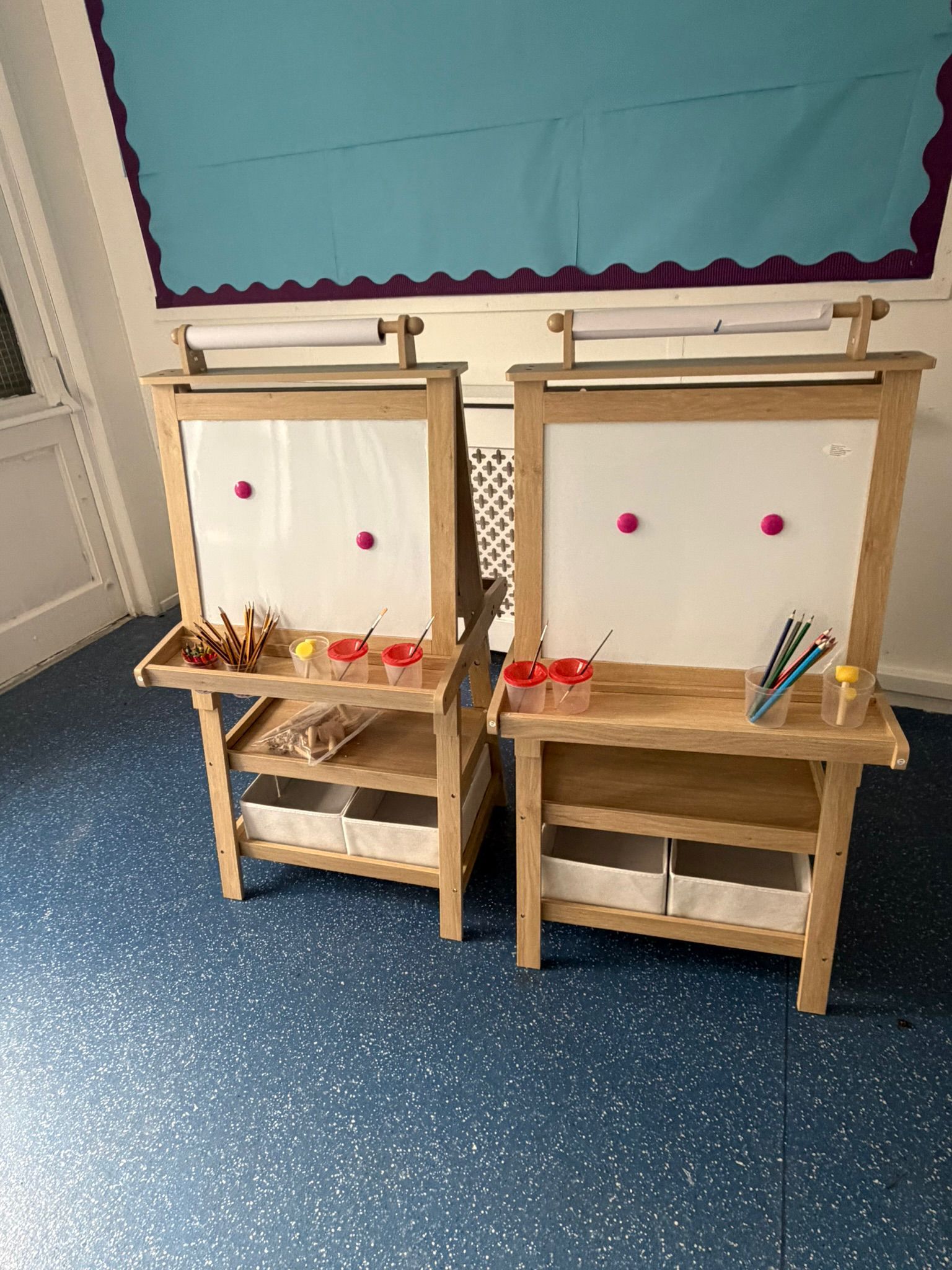 Two wooden children's art easels with whiteboards, paint cups, and supply shelves sit on a blue speckled floor.